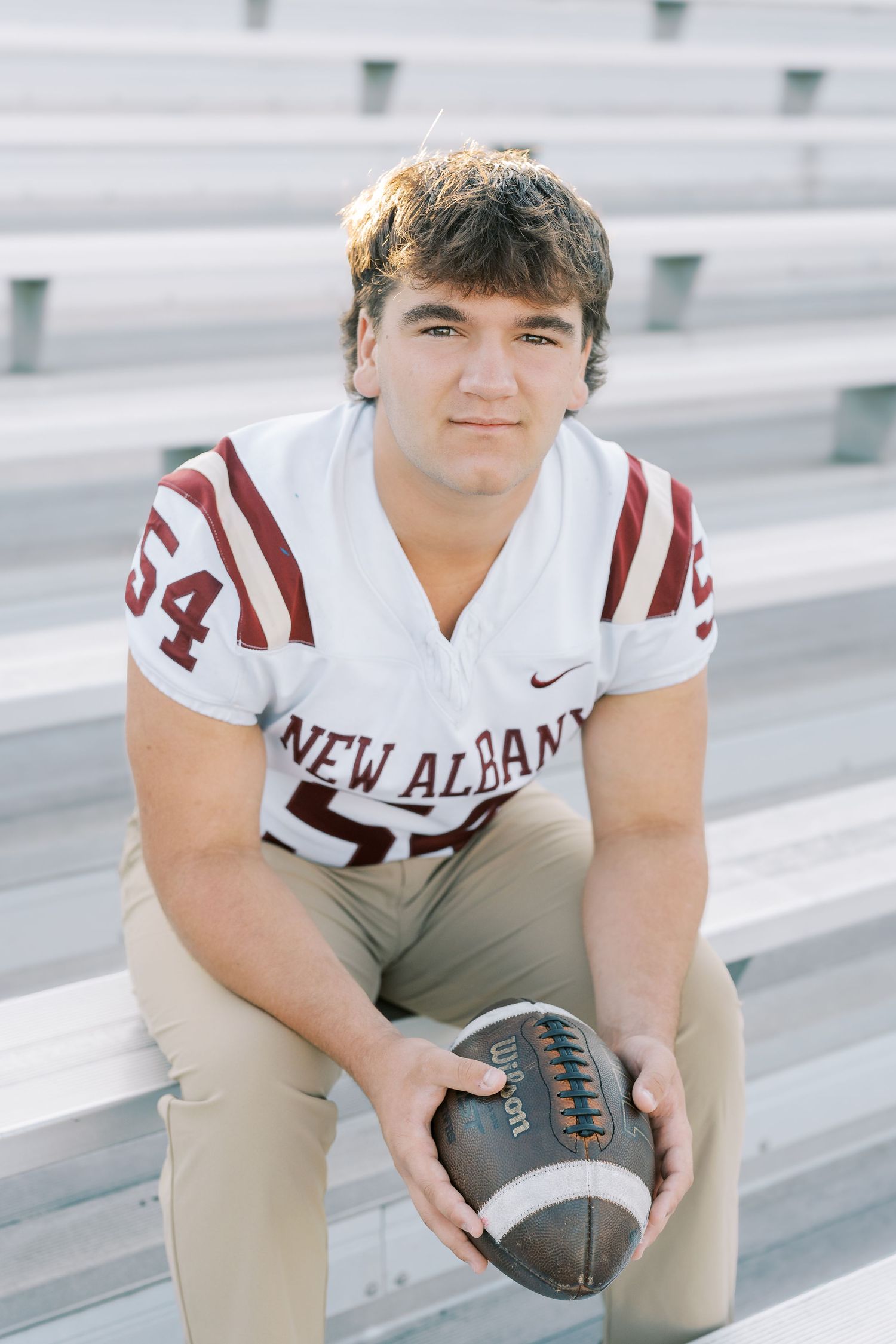 High school football player in white and maroon uniform poses with football on stadium bleachers.