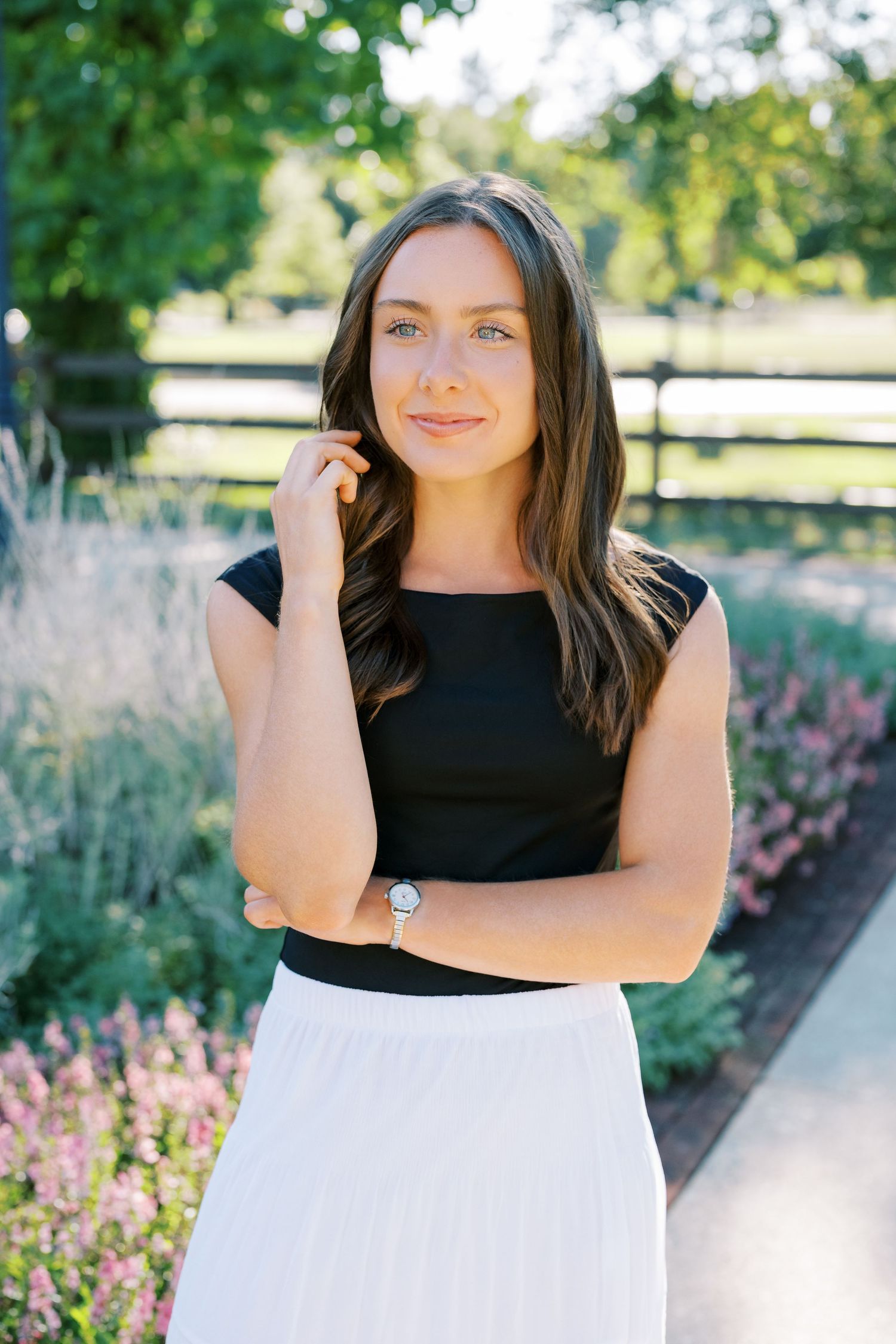 A person in a black and white outfit poses in front of a garden with pink flowers and wooden fencing in the background.