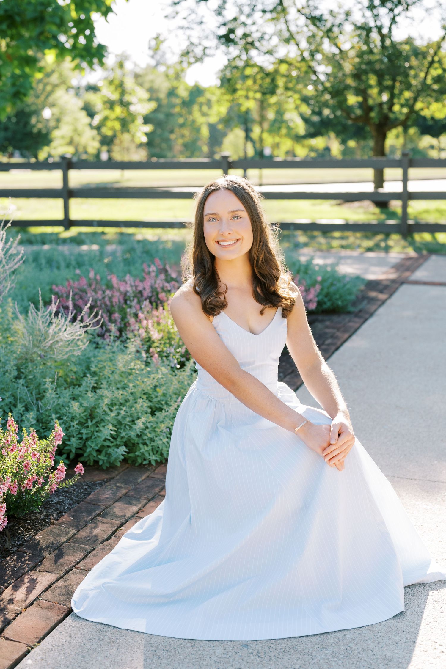 A person in a flowing white dress poses gracefully on garden path surrounded by lush greenery and flowers.
