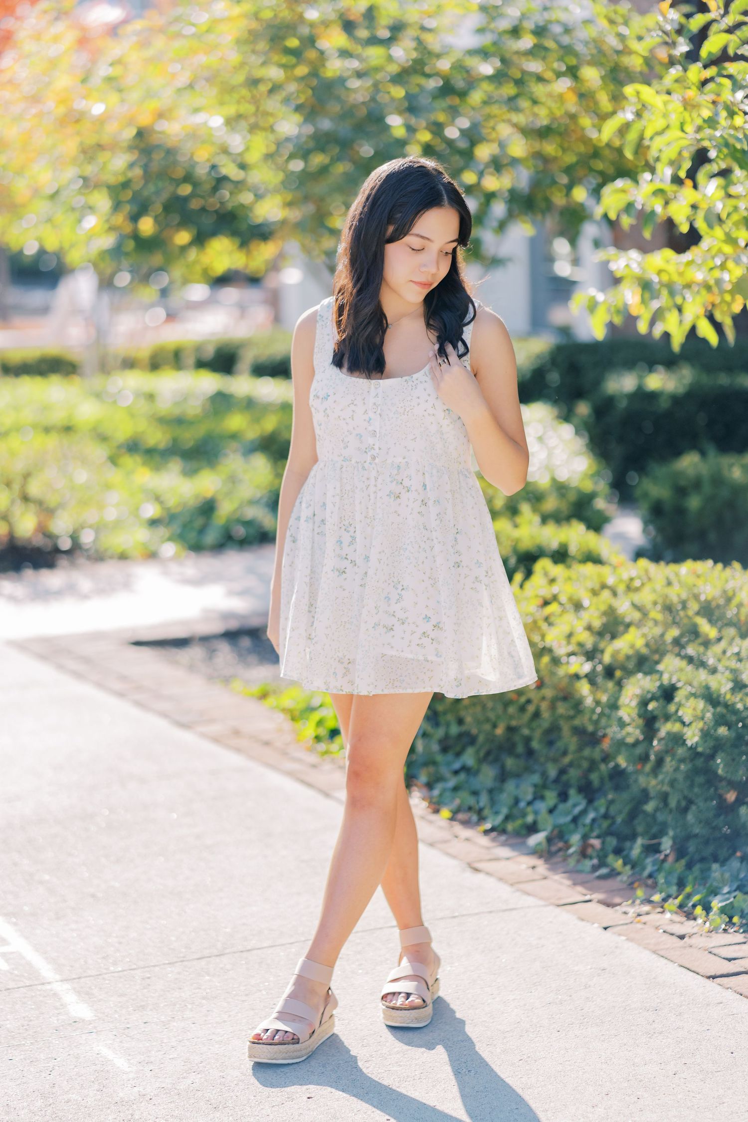 A person in a flowy white summer dress walks along a sunlit pathway with trees and greenery in the background.