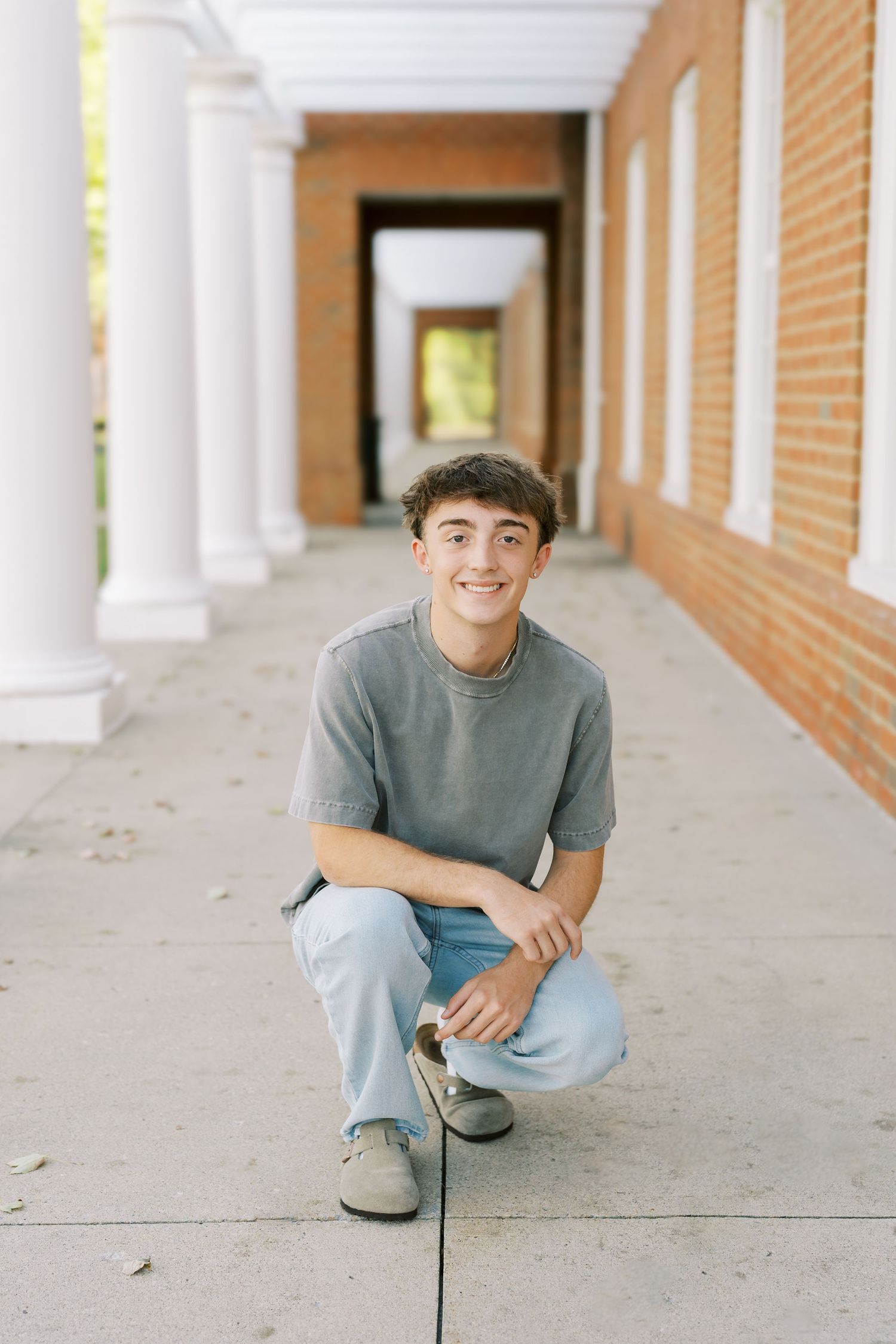 Person in gray t-shirt and light jeans crouches in covered walkway with brick walls and white columns.