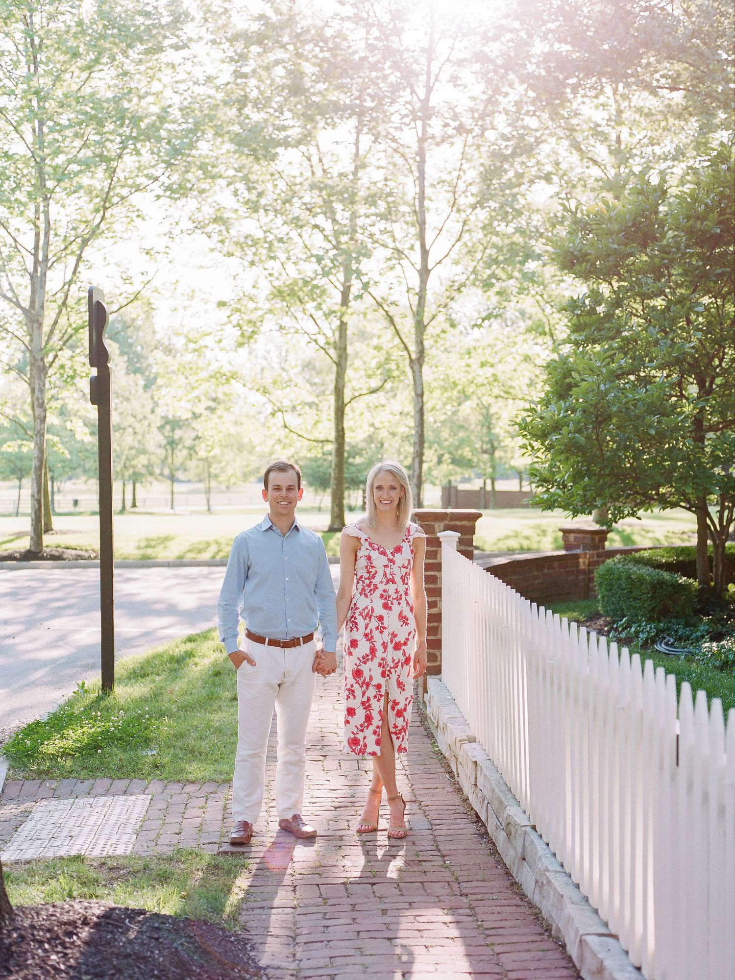 A couple walks along a brick pathway next to a white picket fence on a sunny spring day.