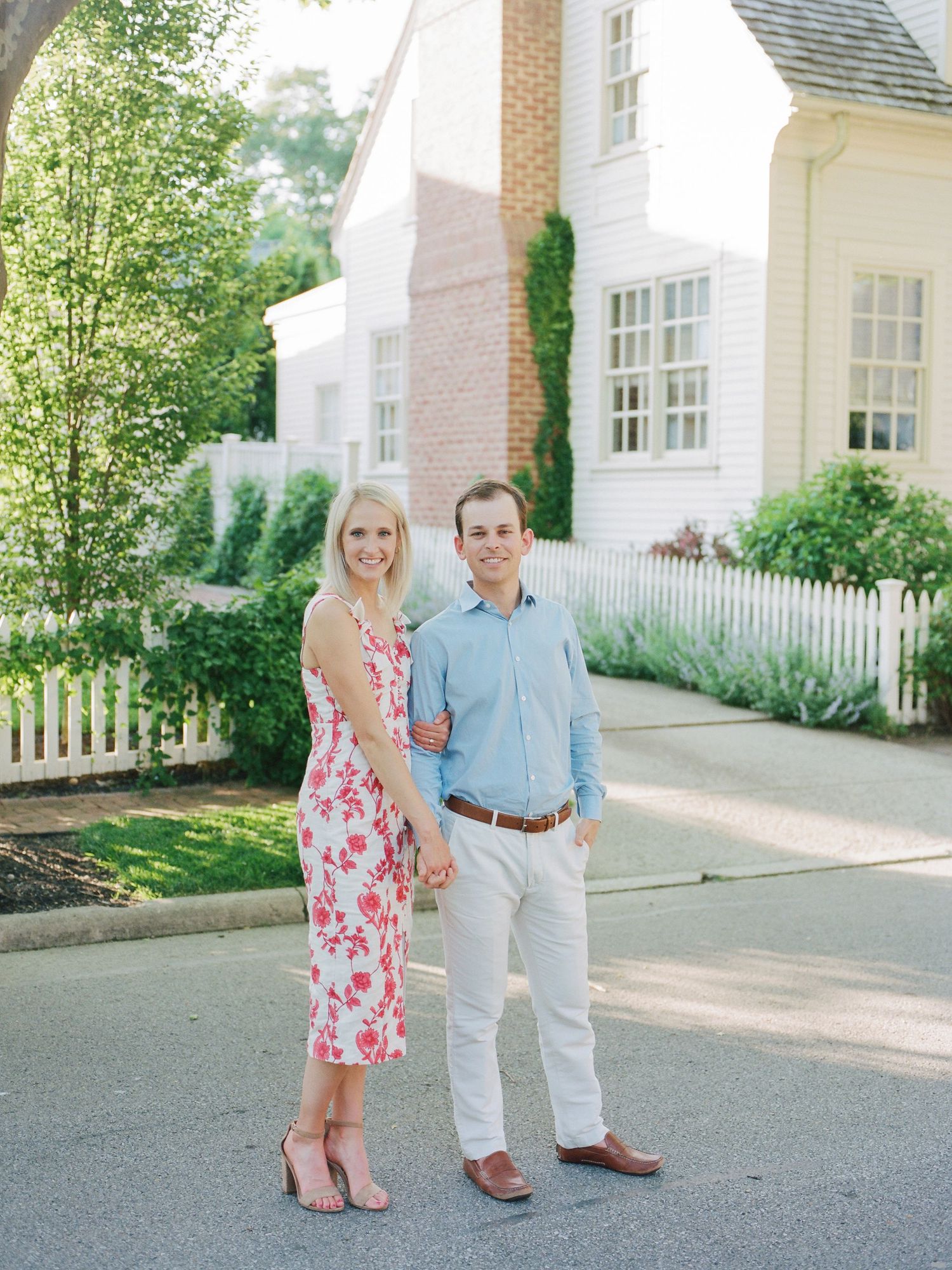 A couple stands together on a paved path next to a white house with a picket fence on a sunny summer day.