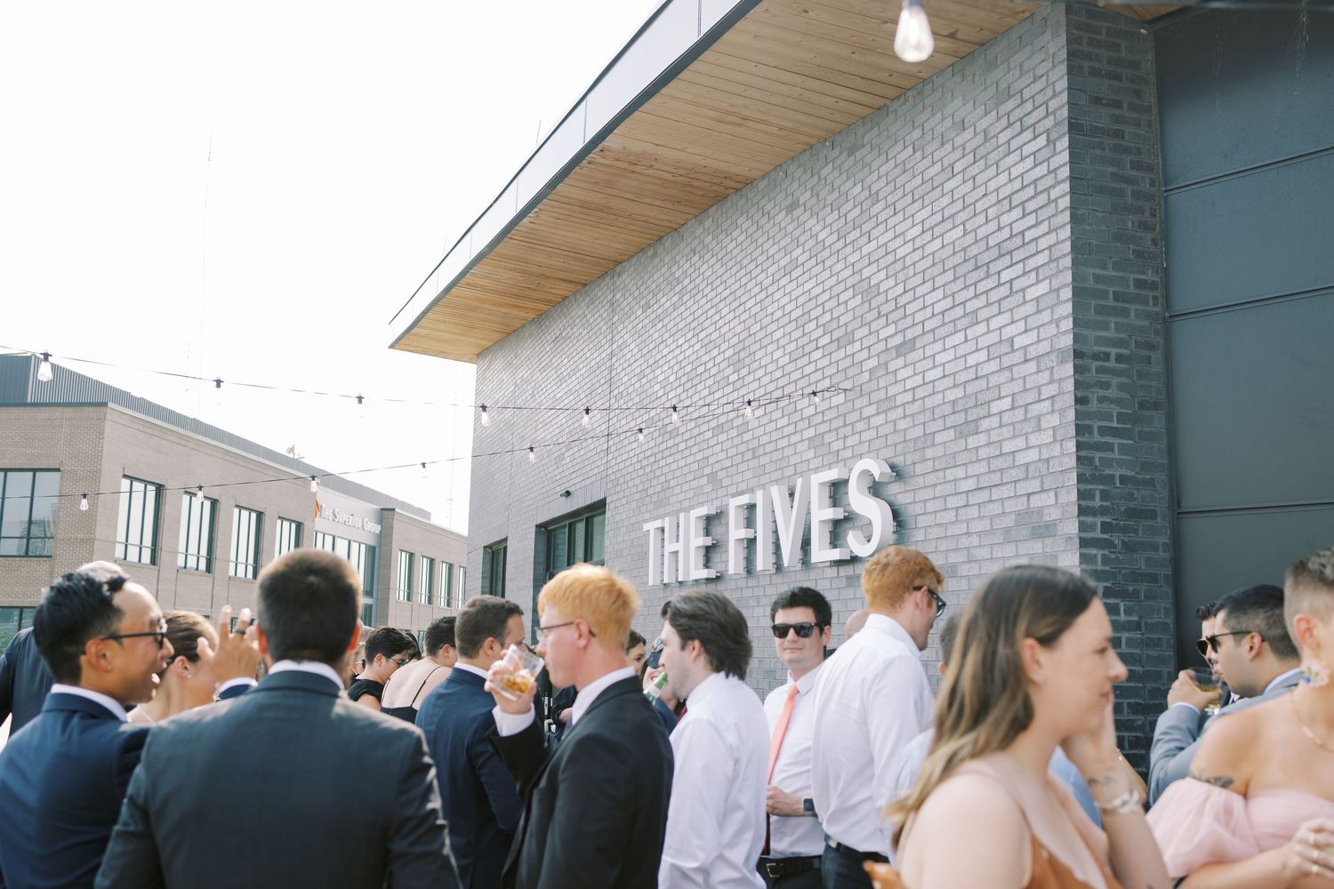 Guests gather outside a brick building entrance during a social event on a sunny day.