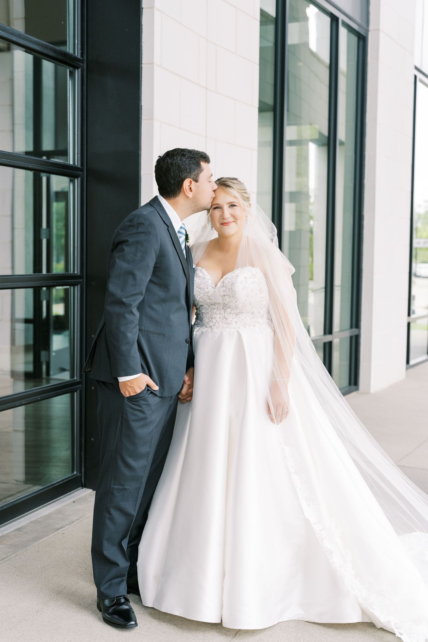 Newlywed couple shares intimate moments in a modern building hallway with floor-to-ceiling windows.