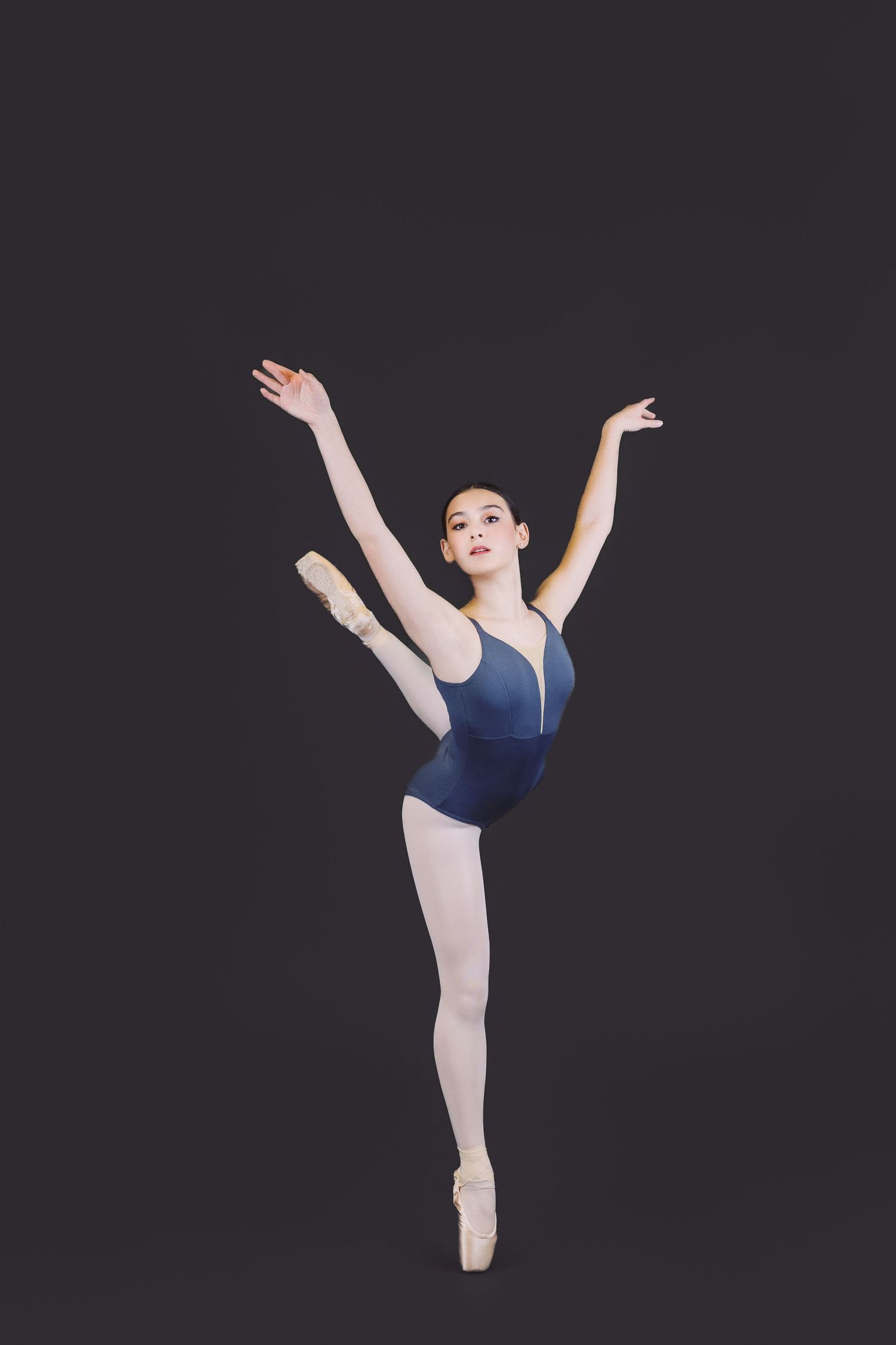 A ballet dancer in a navy blue leotard performs a graceful high kick against a dark background.