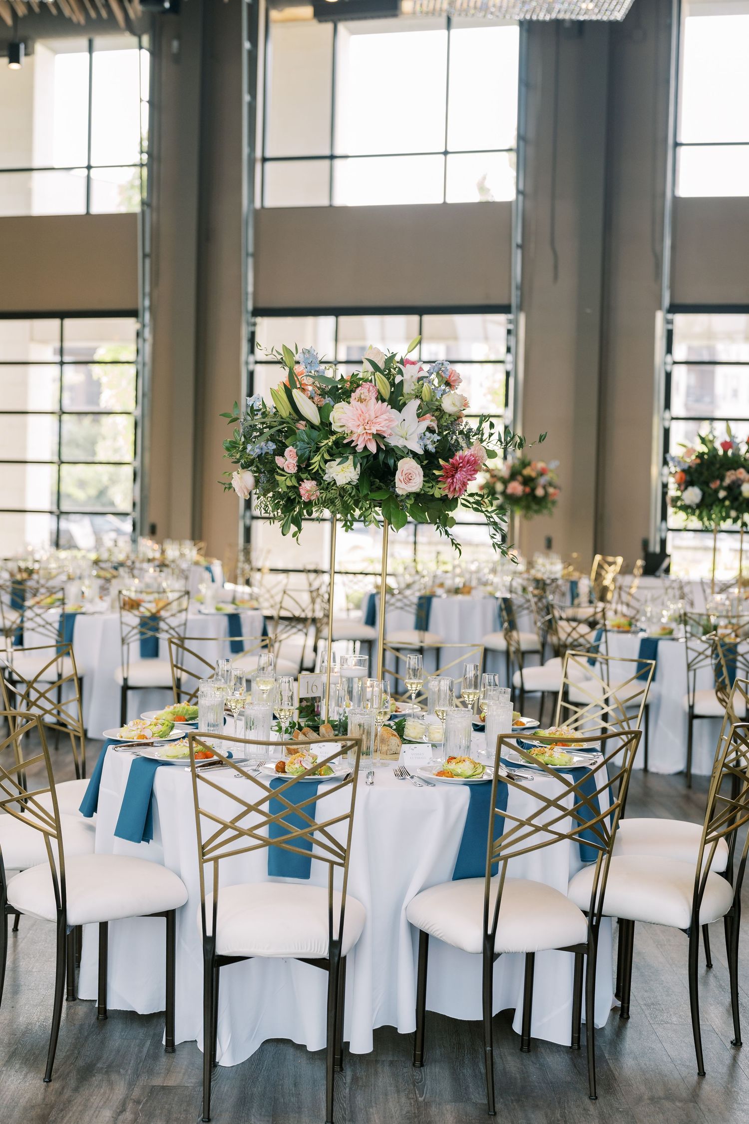 Elegant wedding reception tables with white linens, gold chairs and floral centerpieces in a bright modern venue.