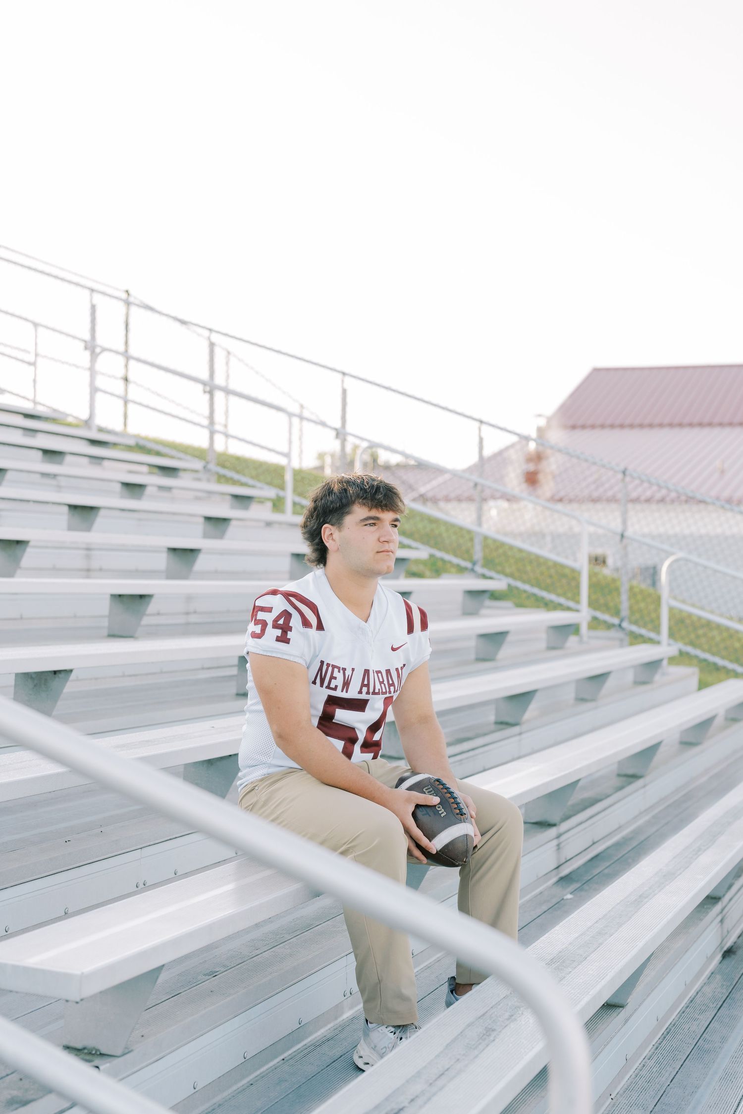 A person in a white jersey sits alone on empty metal bleachers in an outdoor stadium.
