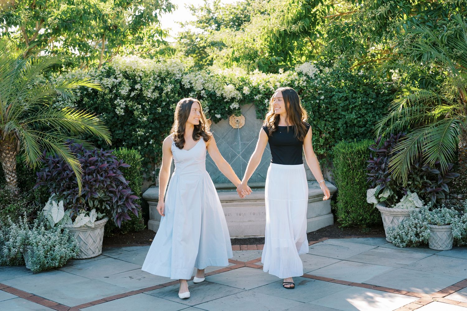 Two people in elegant white dresses hold hands while walking through a lush garden patio setting.