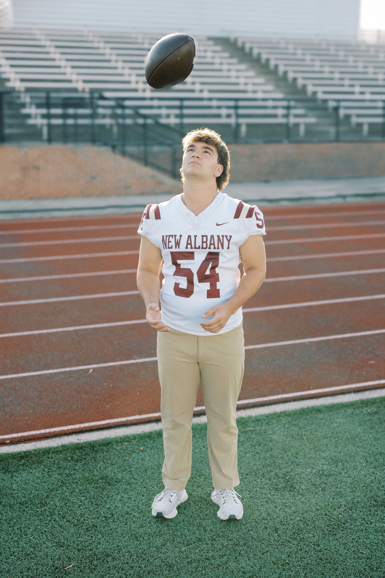 A football player in a New Albany #54 jersey tosses a football in the air on a sports field with empty bleachers.