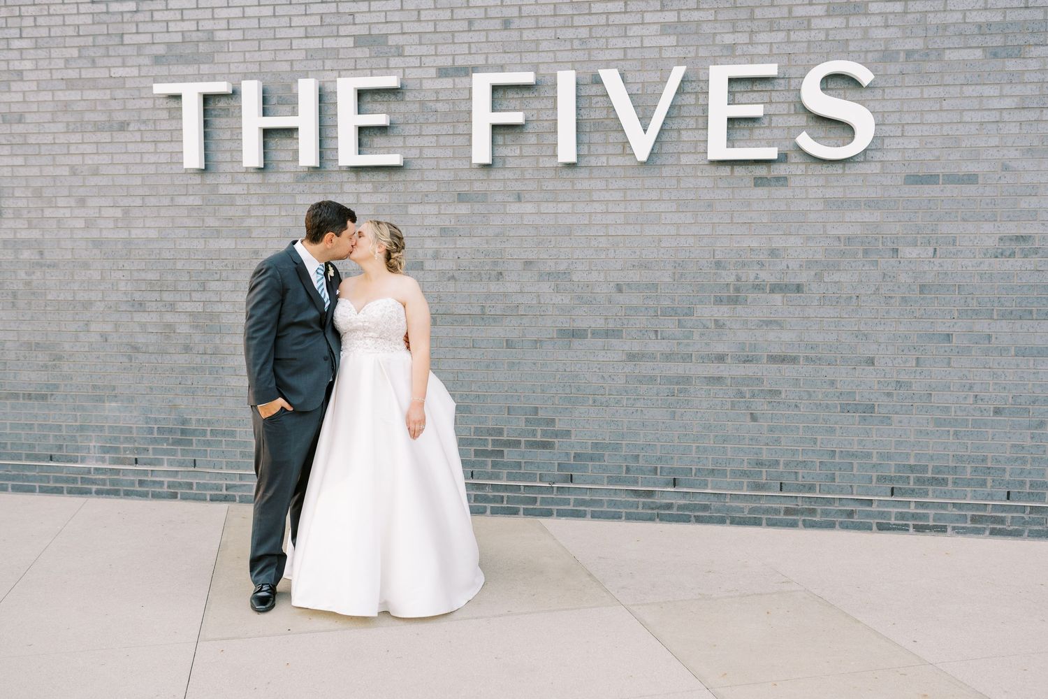 A couple in wedding attire stands against a gray brick wall with 'THE FIVES' text displayed above them.