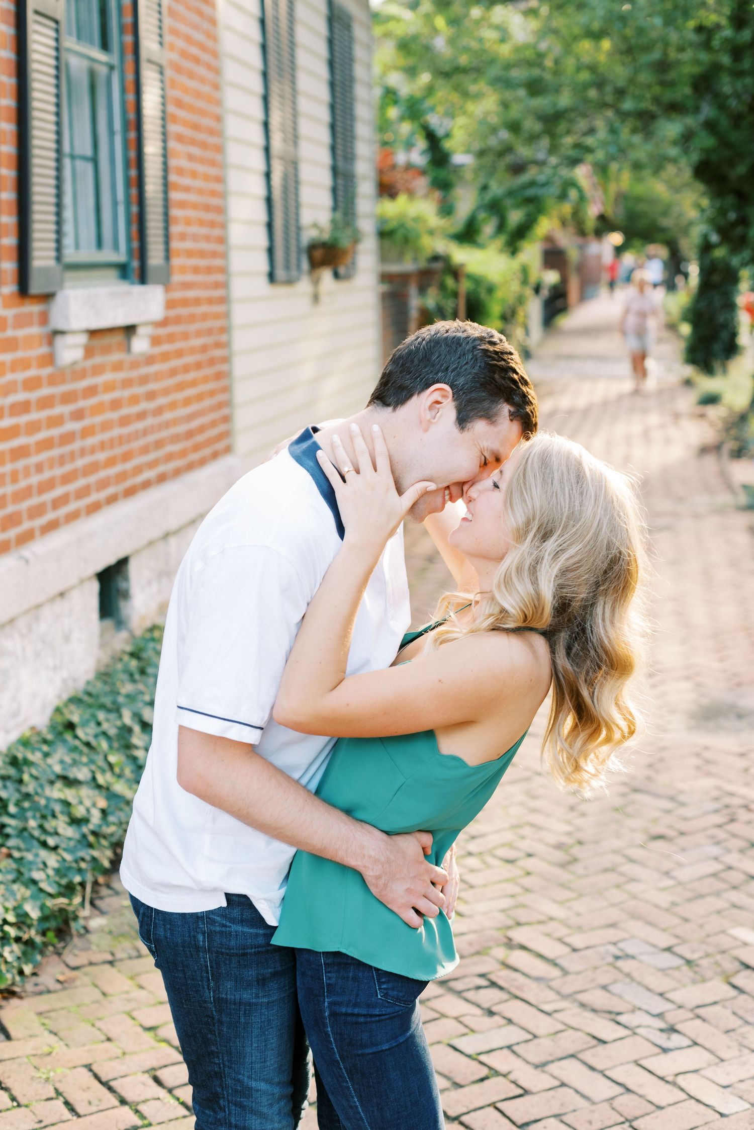 A romantic couple embraces on a brick sidewalk outside a historic building on a sunny afternoon.