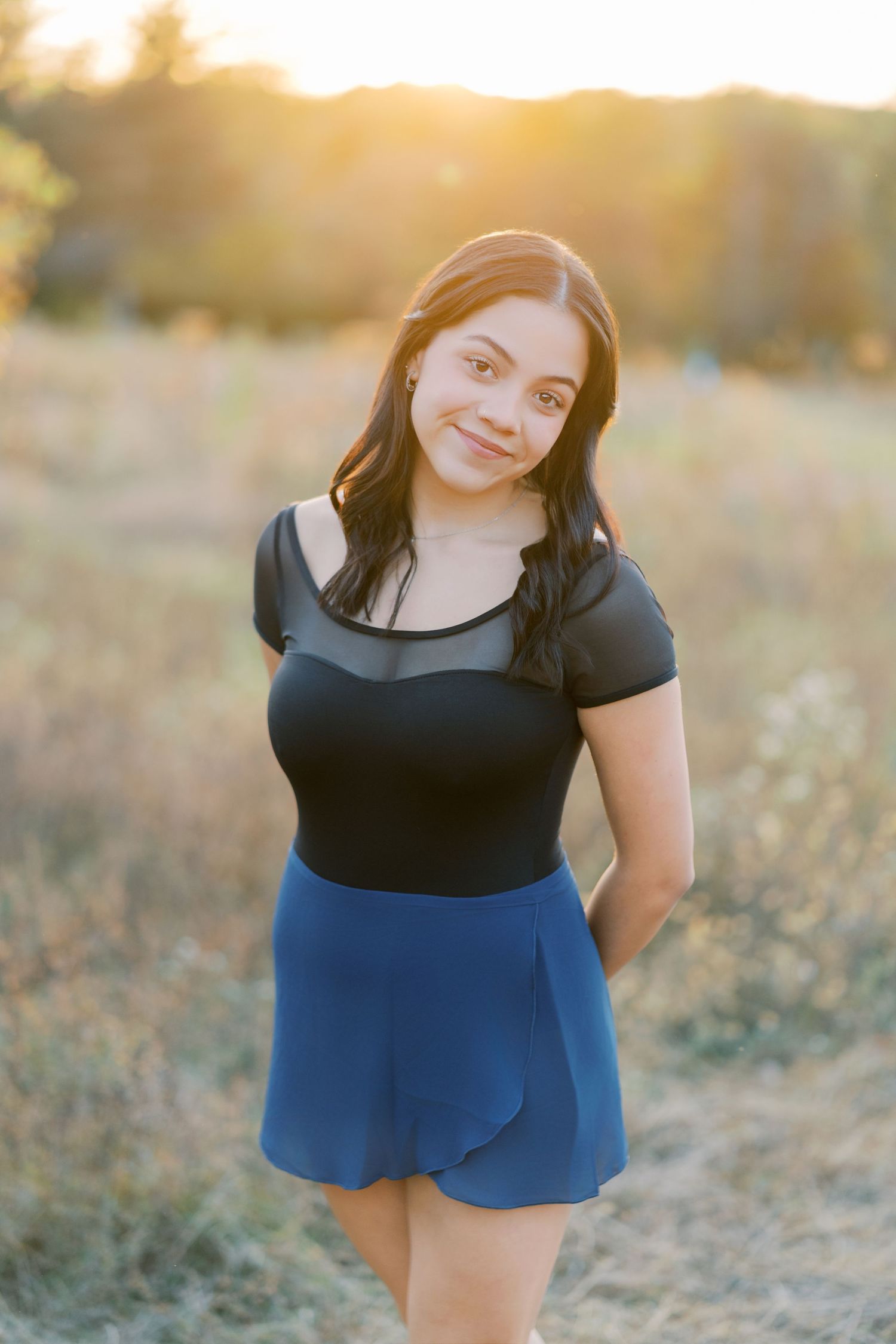 A person wearing a black top and blue skirt poses in a sunlit field during golden hour.