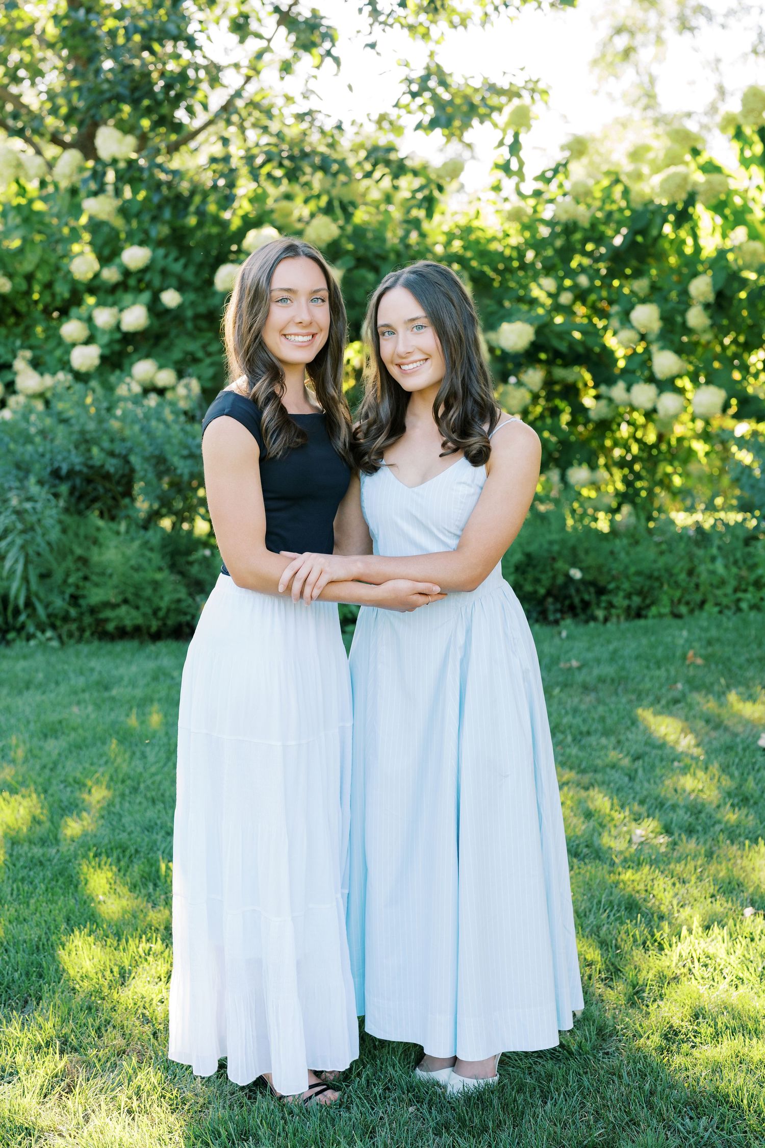 Two people in coordinating black and white dresses pose together in a sunny garden setting with lush greenery.