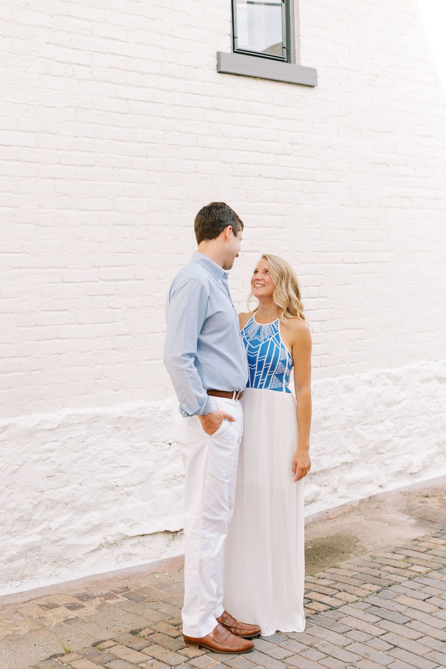 A couple in coordinated white and blue outfits share a romantic moment against a white brick wall on a cobblestone street.