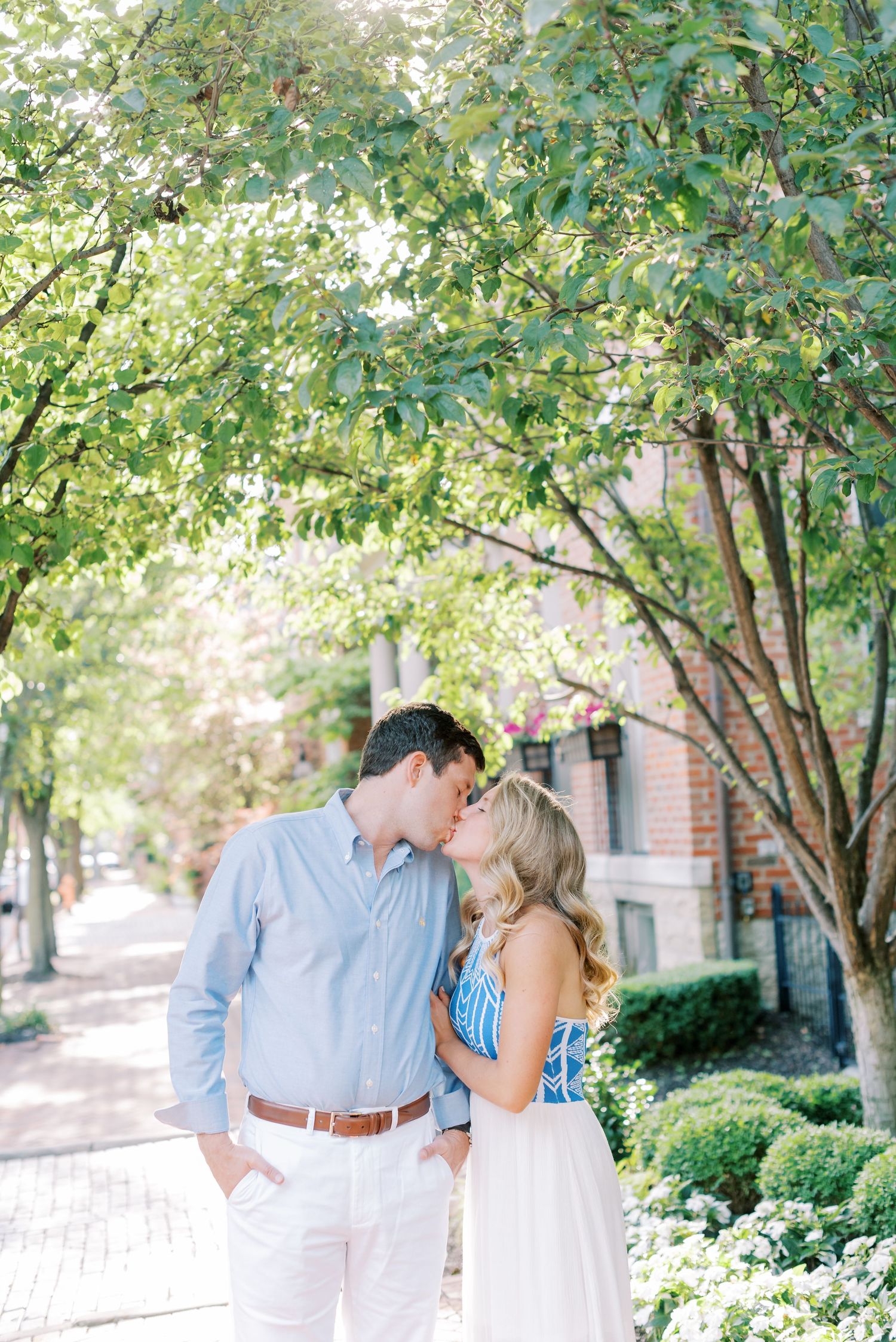 Couple in light blue and white outfits share romantic moments under green trees in a garden setting.