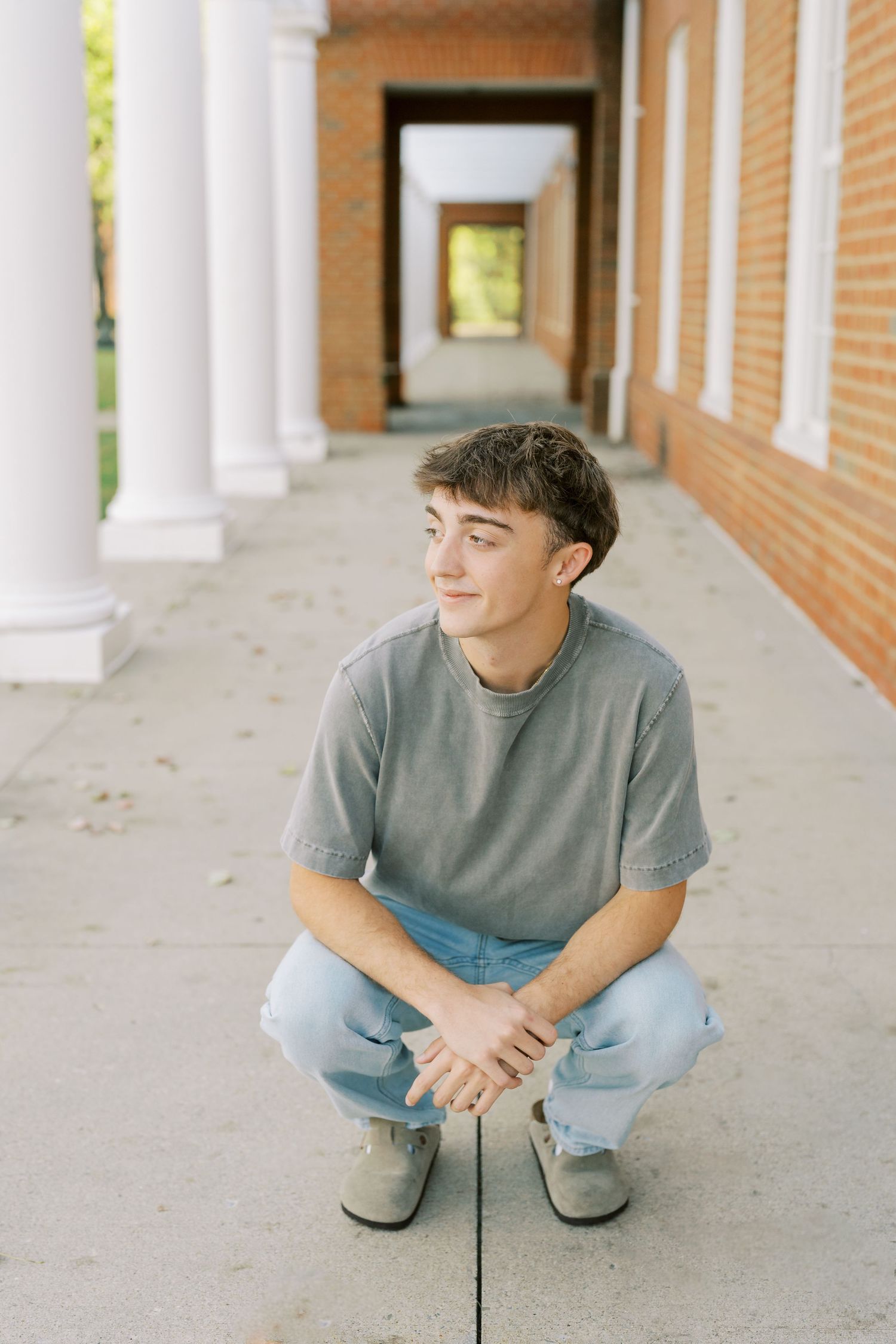A person in casual attire crouches in a covered walkway with brick walls and white columns.