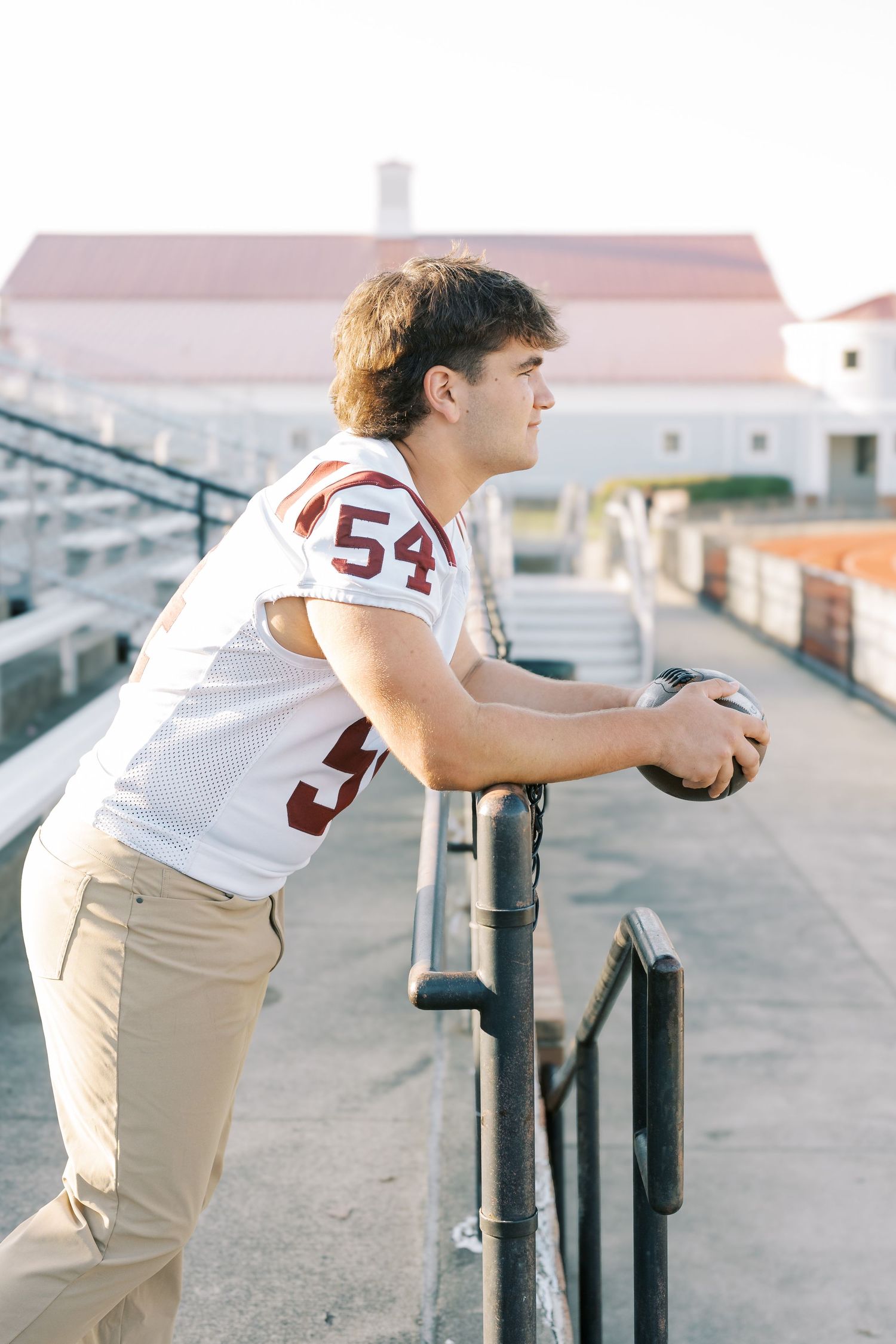 Athletic person in white jersey and khaki pants leans against metal railing at sunset.