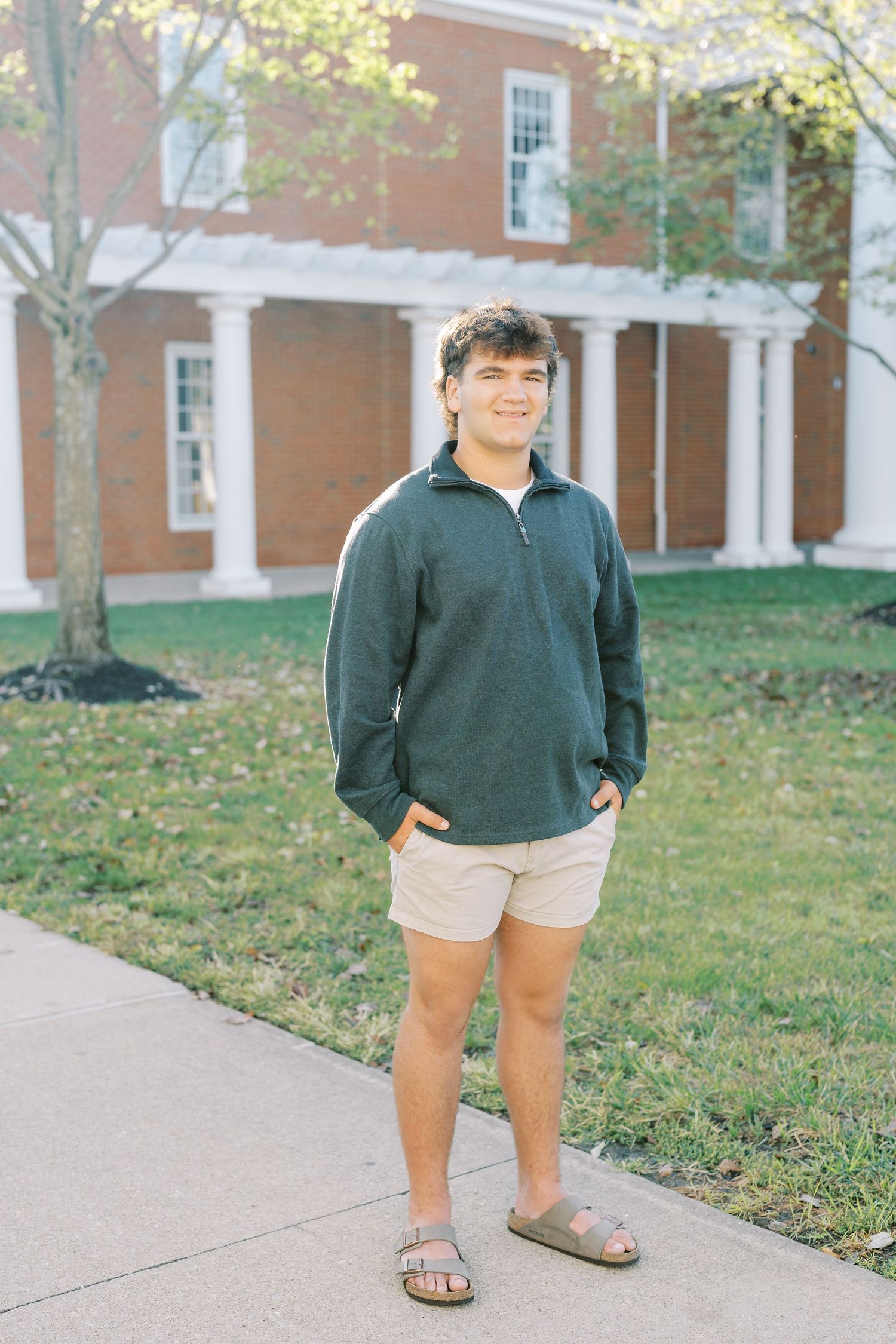A person in a green sweater and khaki shorts stands on a sidewalk near brick buildings on a sunny autumn day.