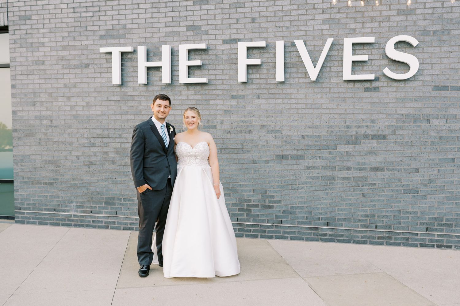 A wedding couple poses against a gray brick wall with text reading 'THE FIVES' mounted on the surface.