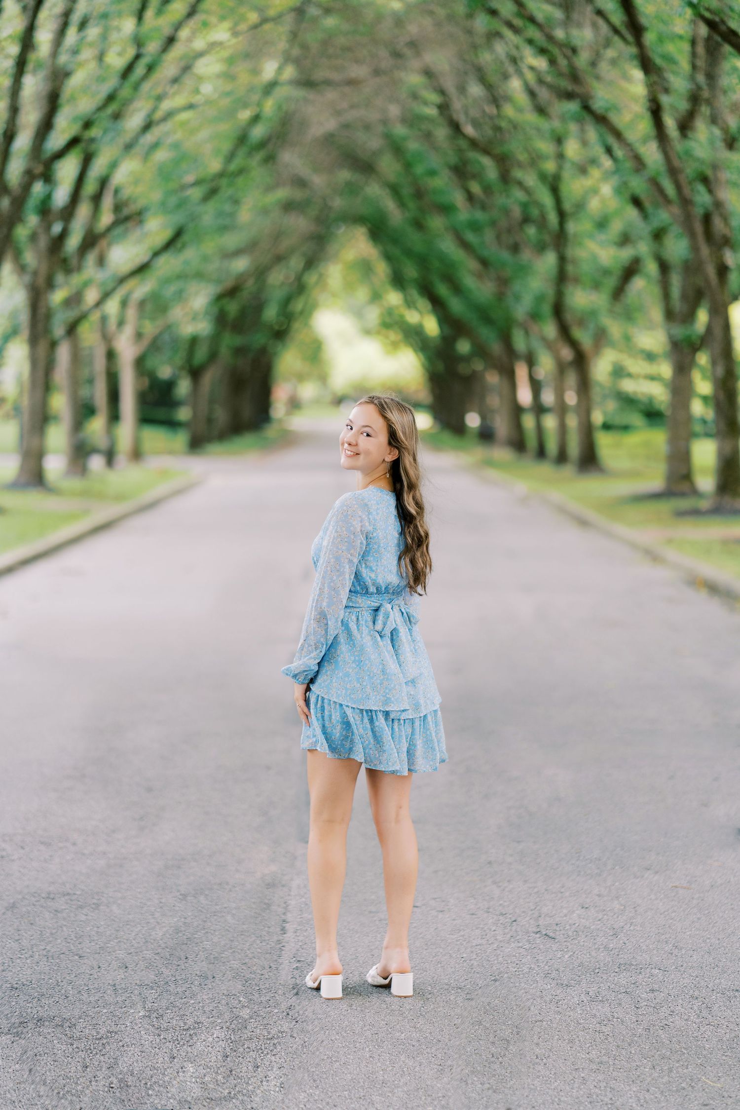 A person in a light blue ruffled dress walks down a tree-lined path on a bright summer day.