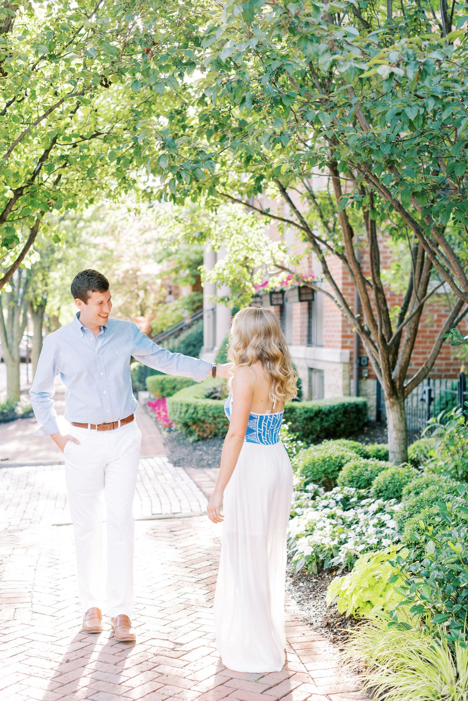 Couple walking and holding hands along a brick path surrounded by lush green landscaping on a sunny day.