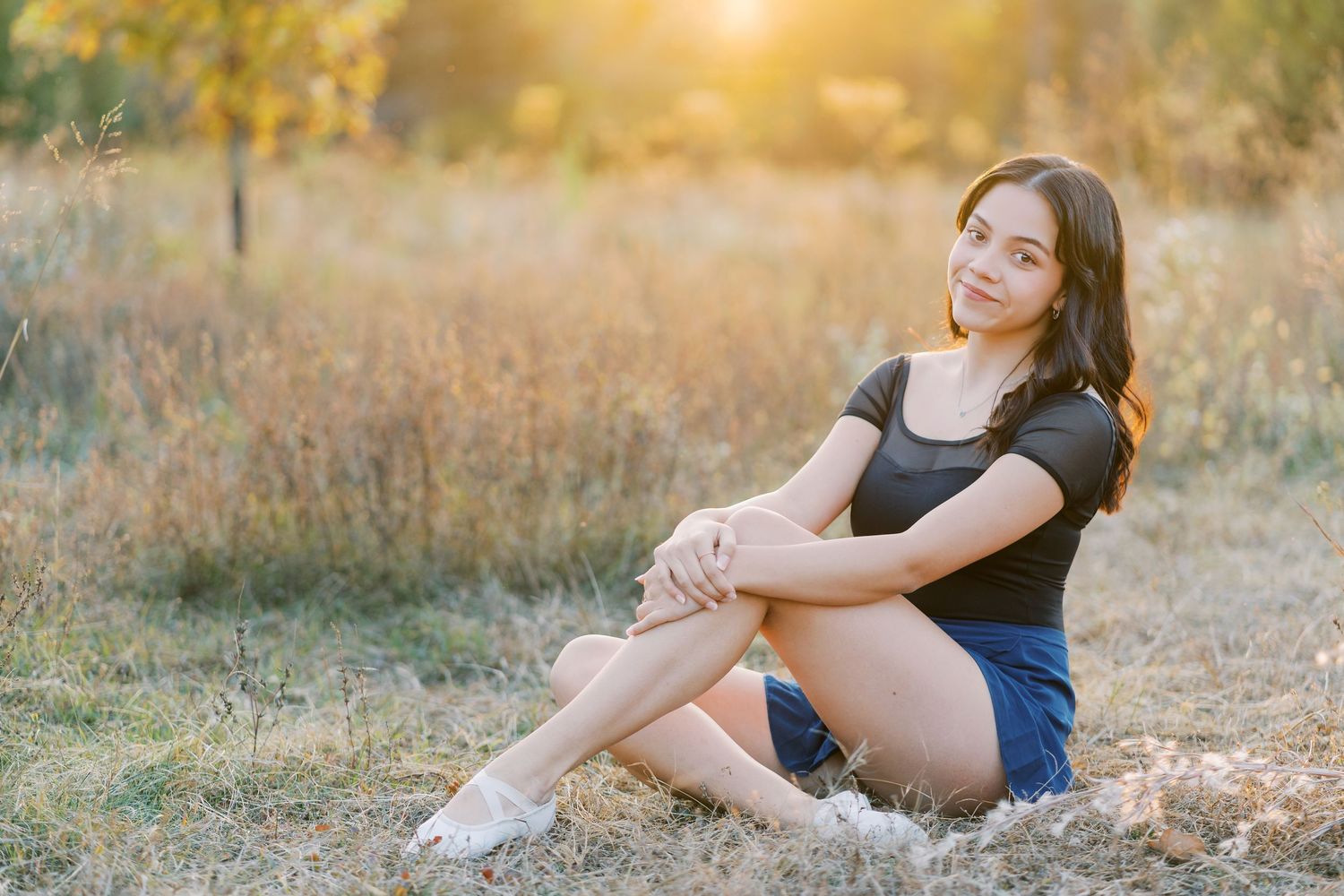 A person sits in a sunlit field wearing a navy dress during golden hour at sunset.