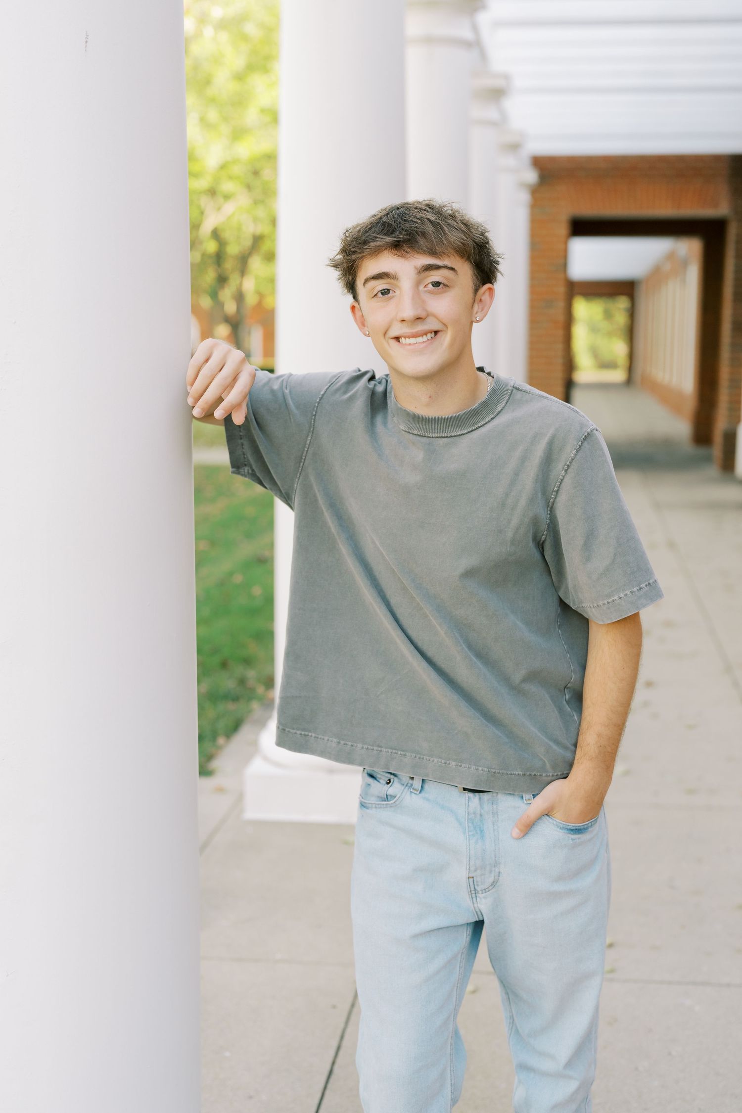 A young person in a gray t-shirt and light jeans poses casually against white pillars outdoors.