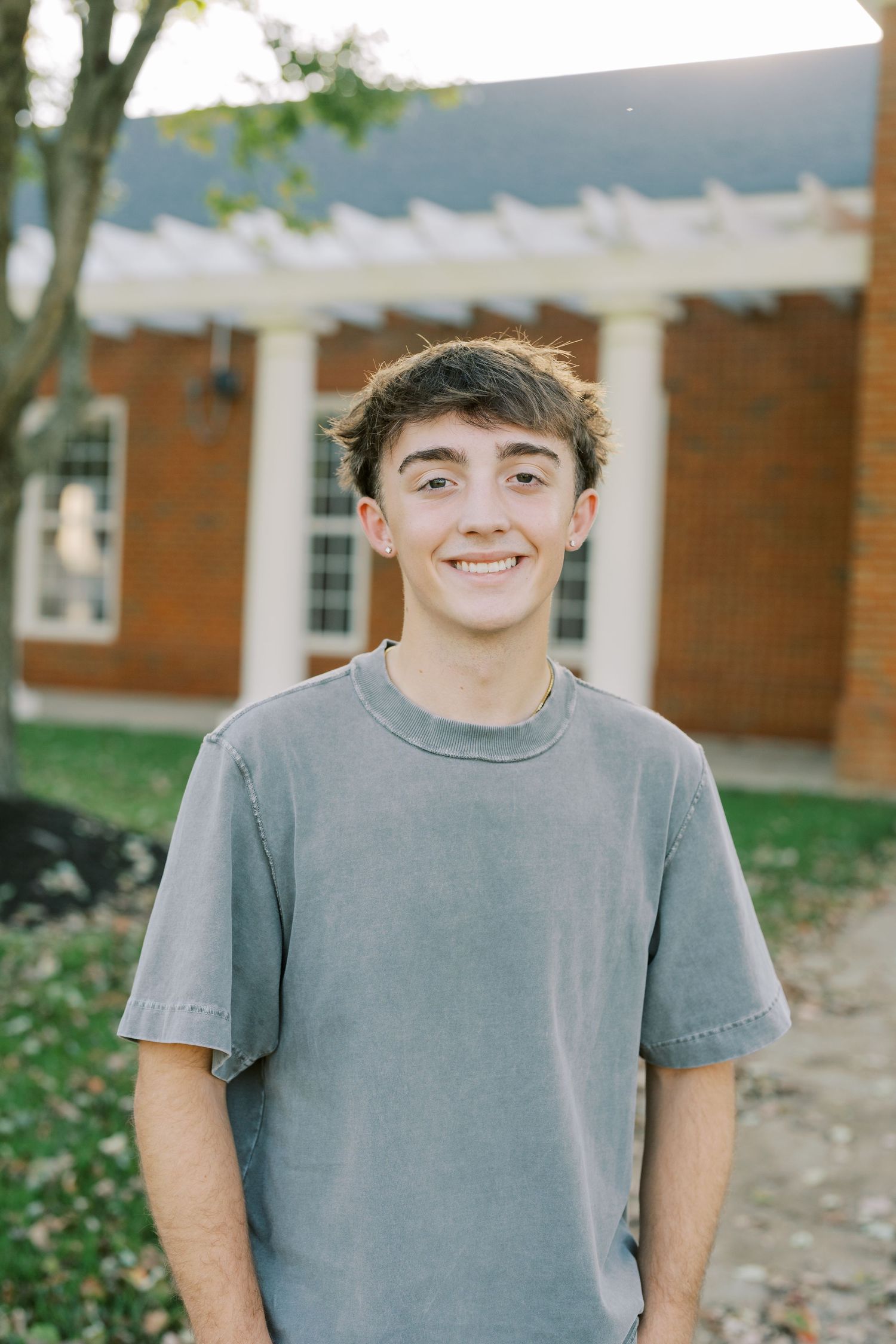 A person in a grey t-shirt stands smiling outdoors in front of a brick building with a white pergola.