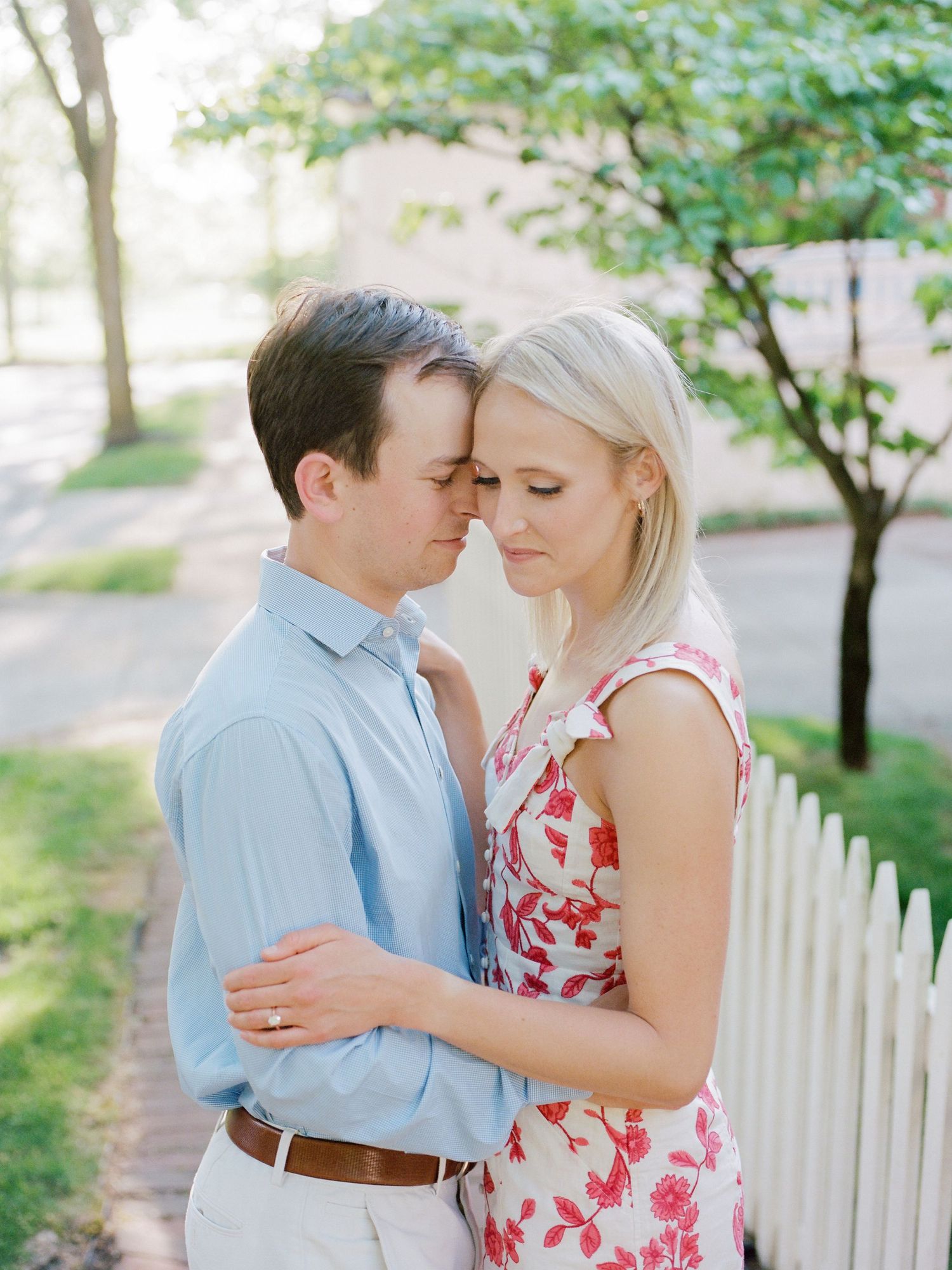 Romantic couple in light blue shirt and floral dress embrace tenderly by white picket fence on spring day.