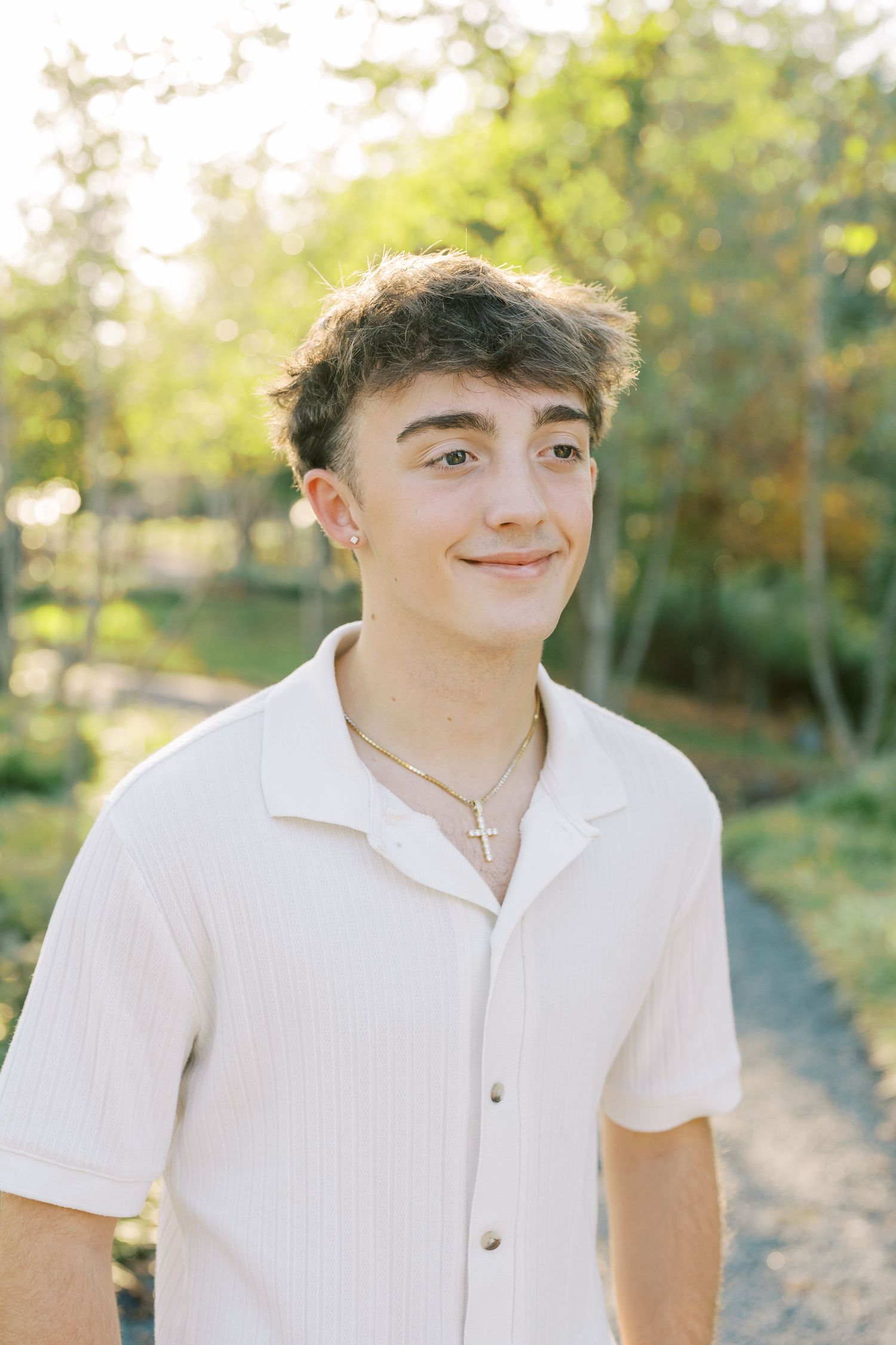 A person wearing a white button-down shirt poses outdoors in a natural setting with sunlit trees in the background.