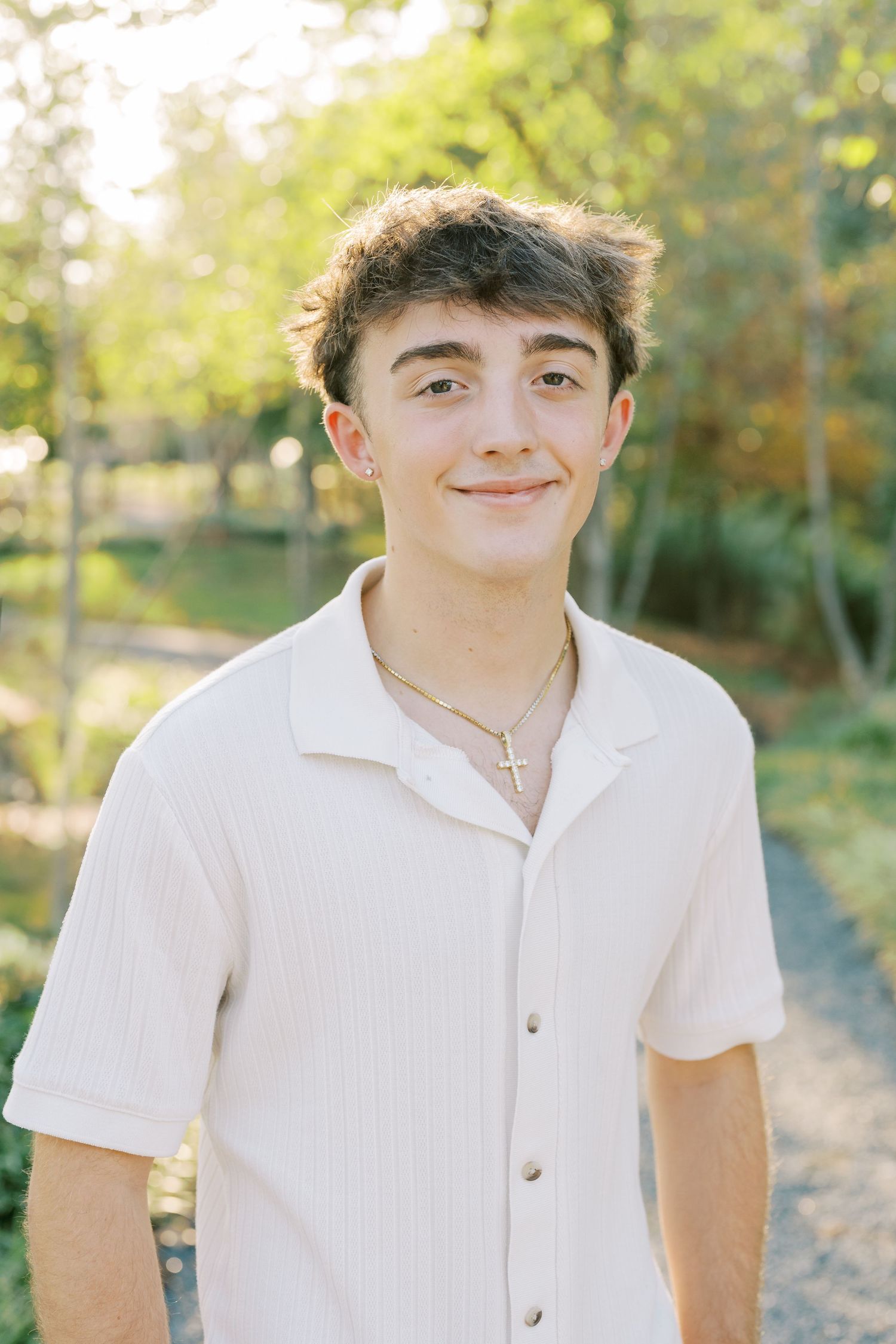 Young person wearing a white polo shirt poses outdoors in natural lighting with trees and foliage in the background.