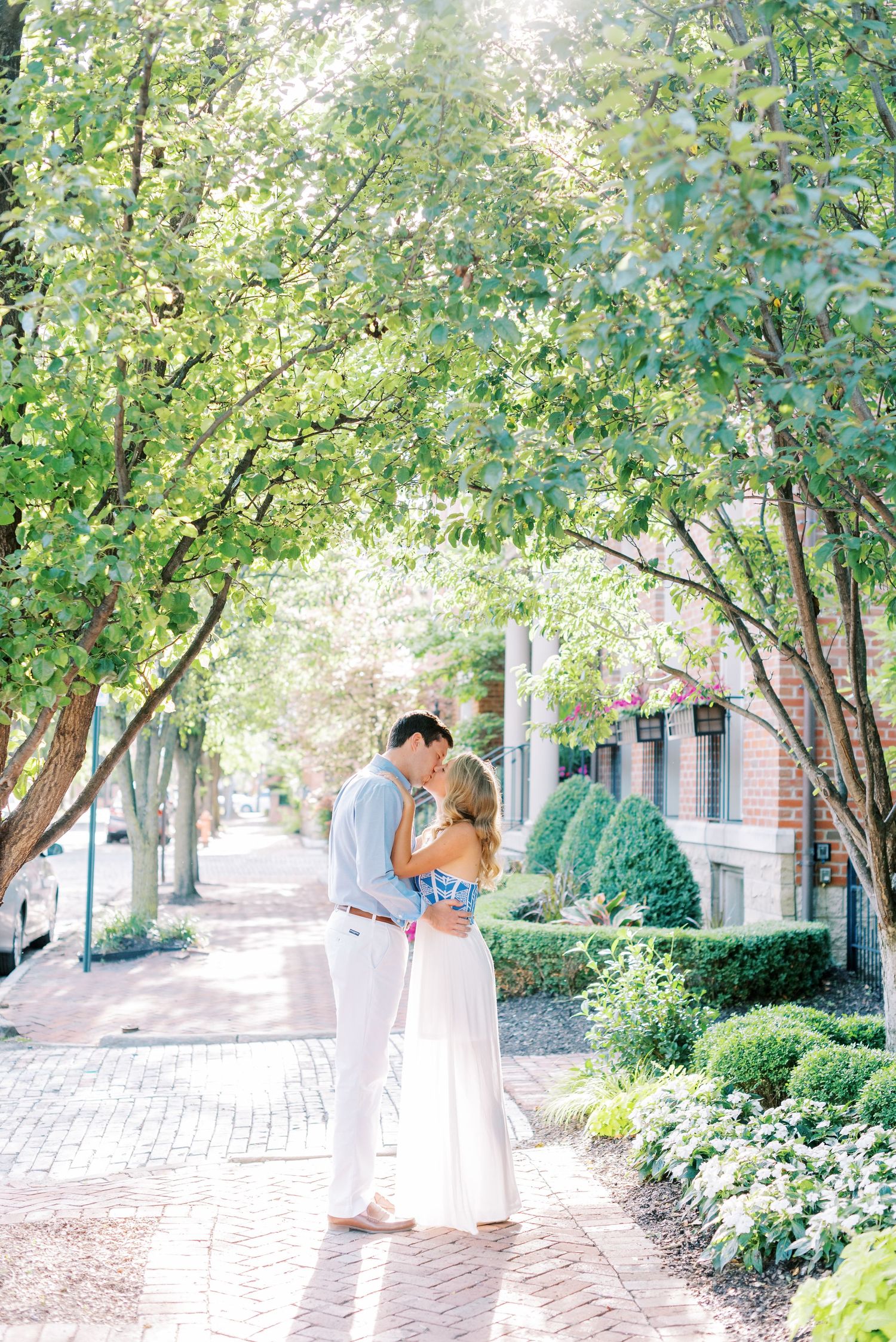 A couple shares a romantic moment under green trees along a brick pathway on a bright sunny day.