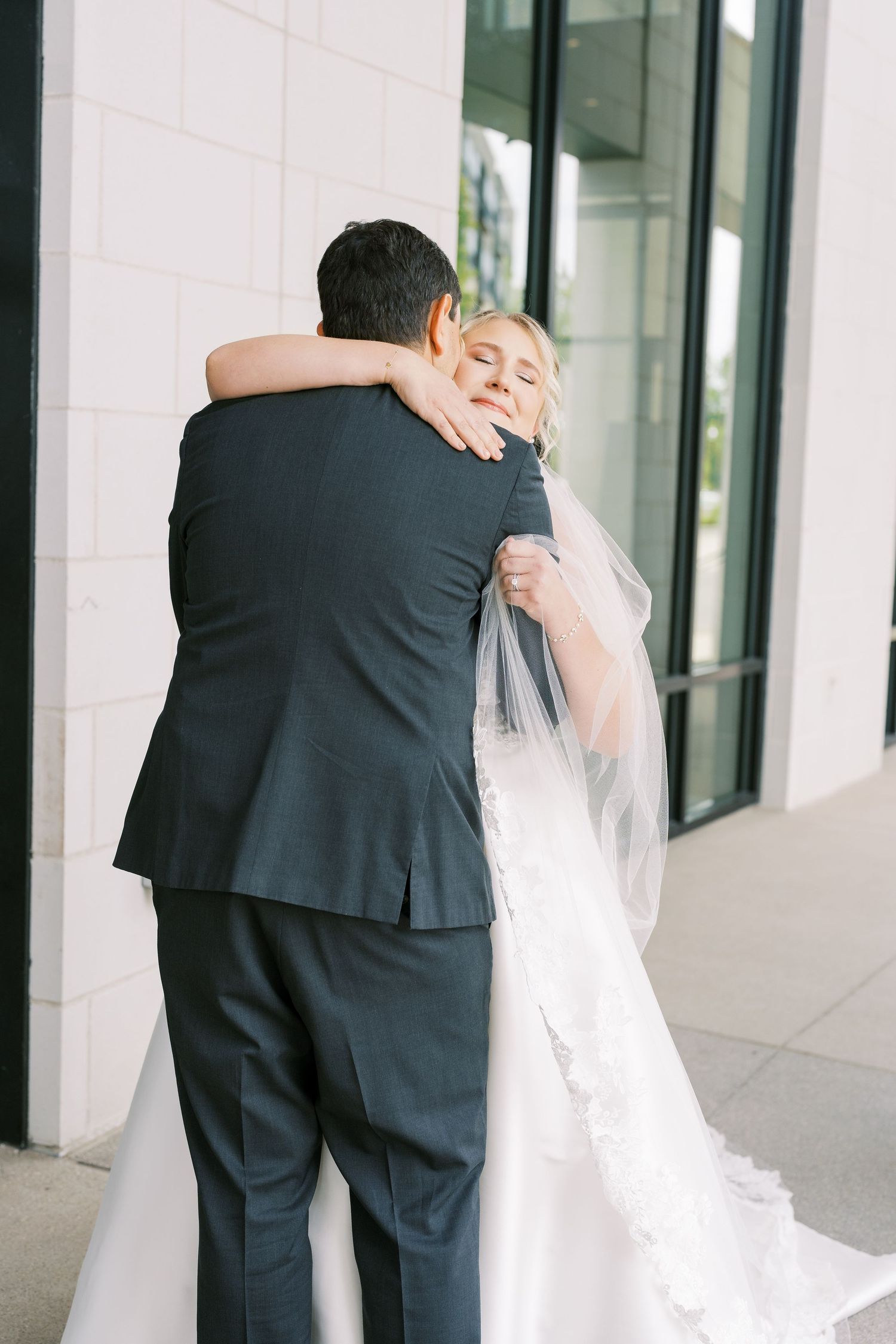 Bride in flowing white gown embraces groom in black suit during intimate wedding moment against modern building exterior.
