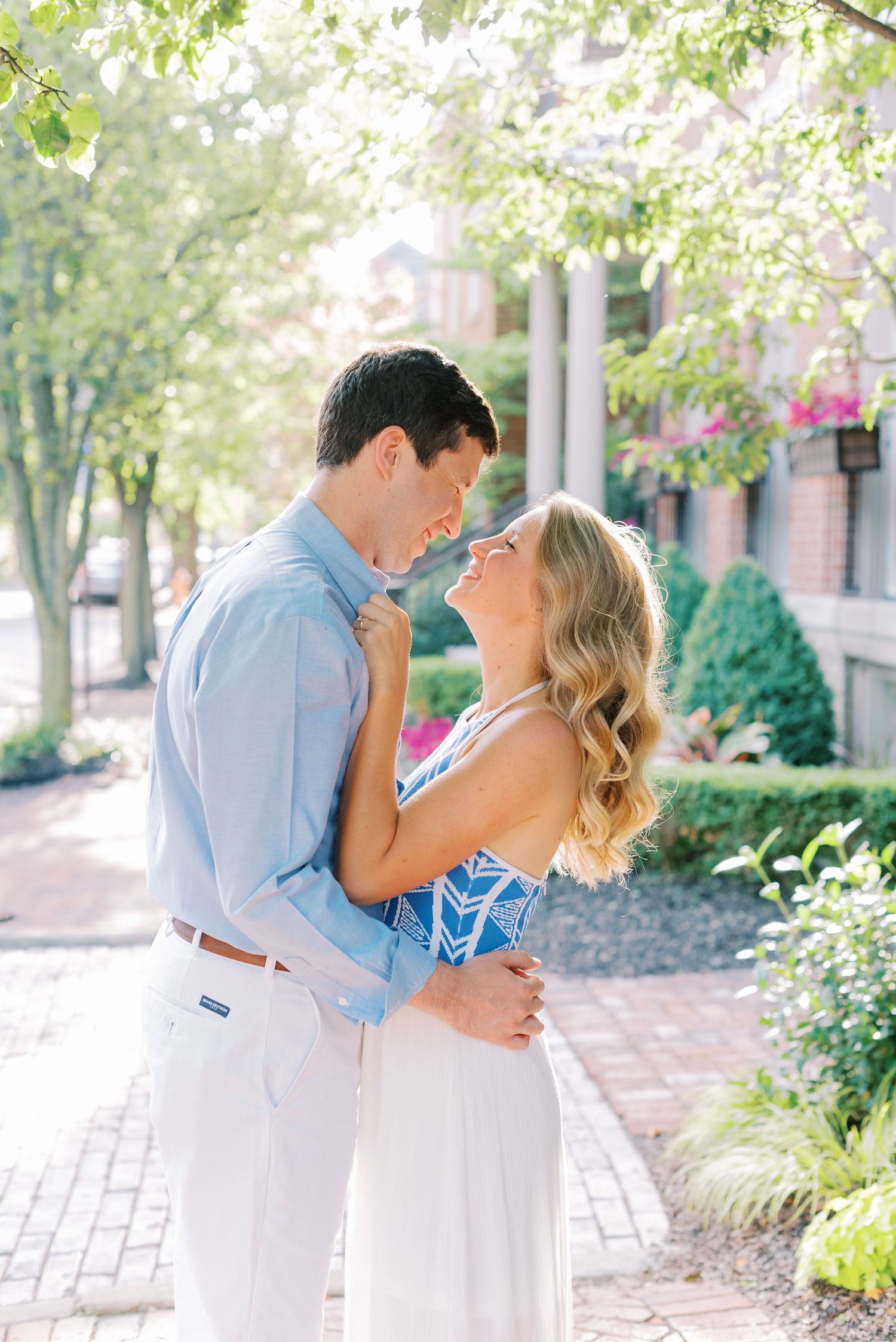 A romantic couple in coordinating blue and white outfits share tender moments on a brick pathway surrounded by greenery.