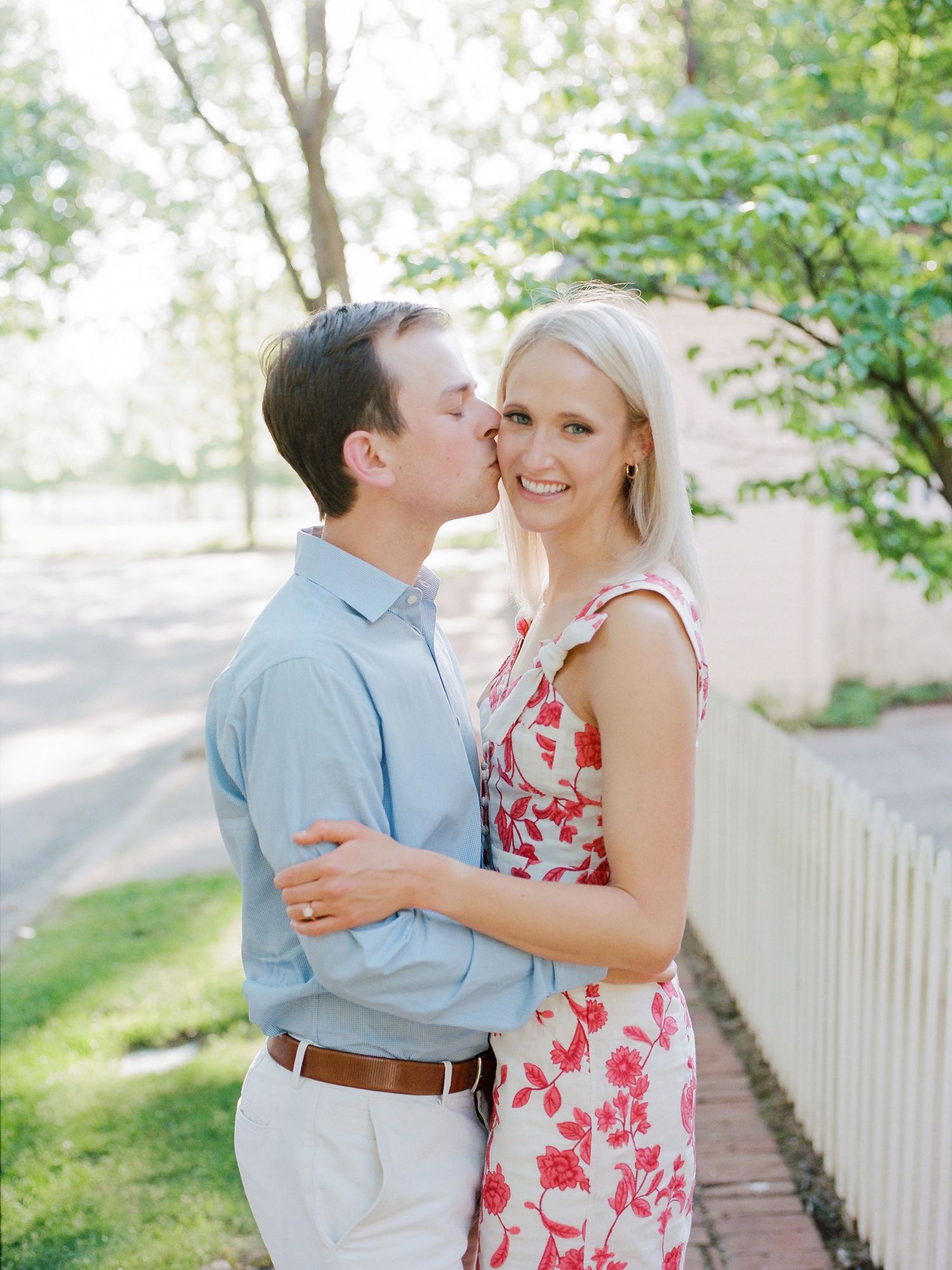 A couple embraces on a sunny path with a white picket fence and green foliage in the background.