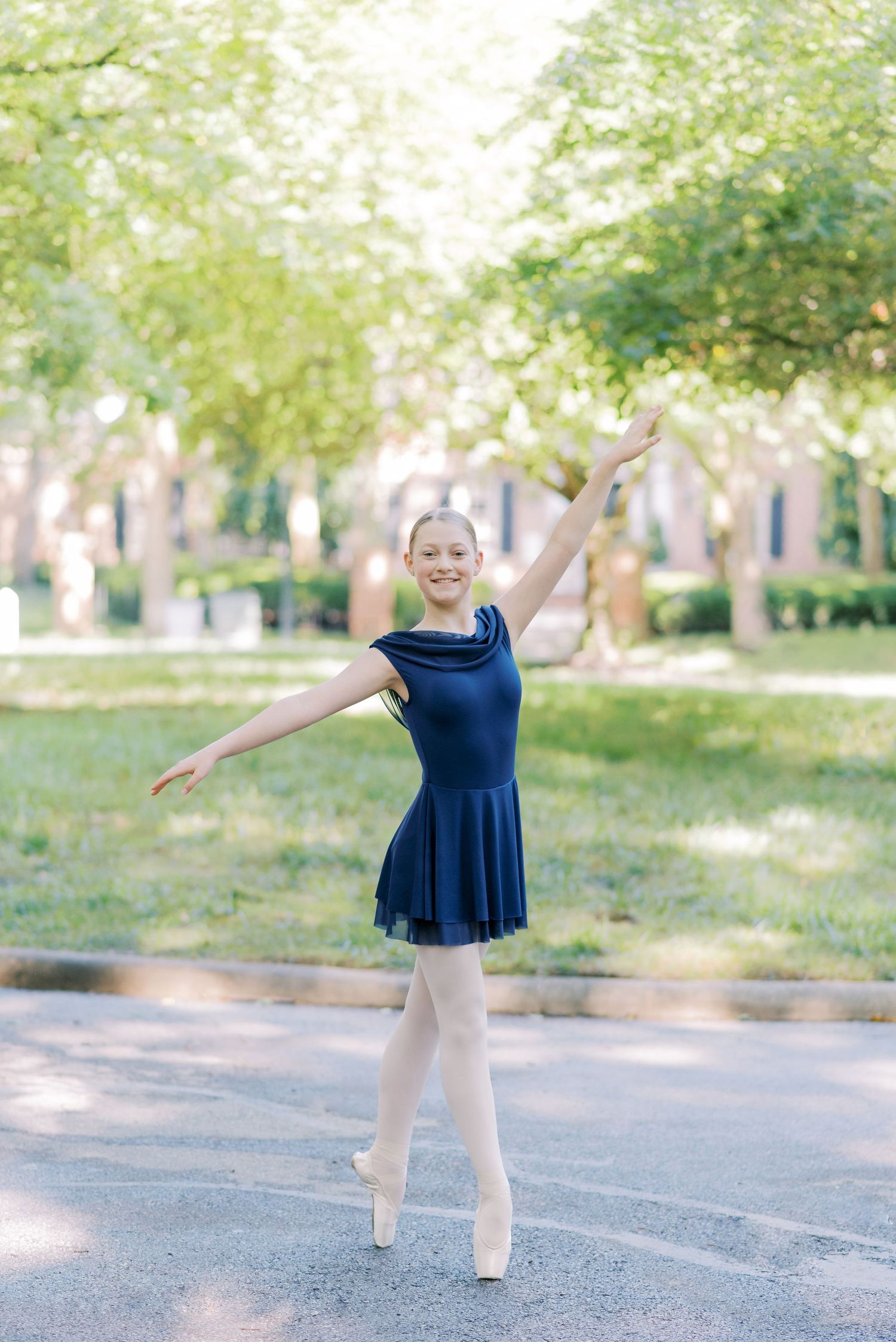 A ballet dancer in a navy blue dress performs en pointe on a tree-lined path in soft natural lighting.