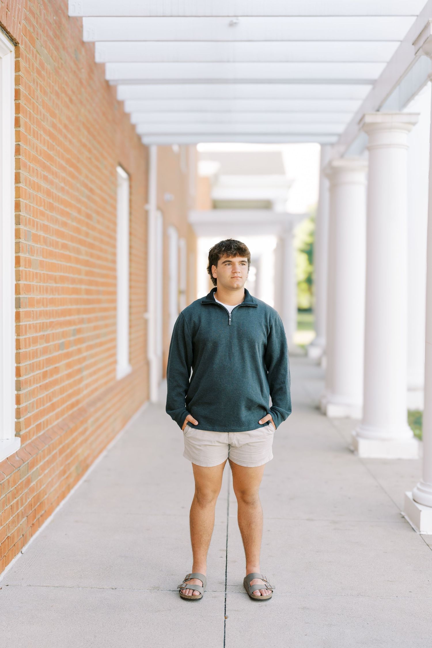 A person stands in a covered walkway wearing a green sweater and khaki shorts with white columns and brick walls.