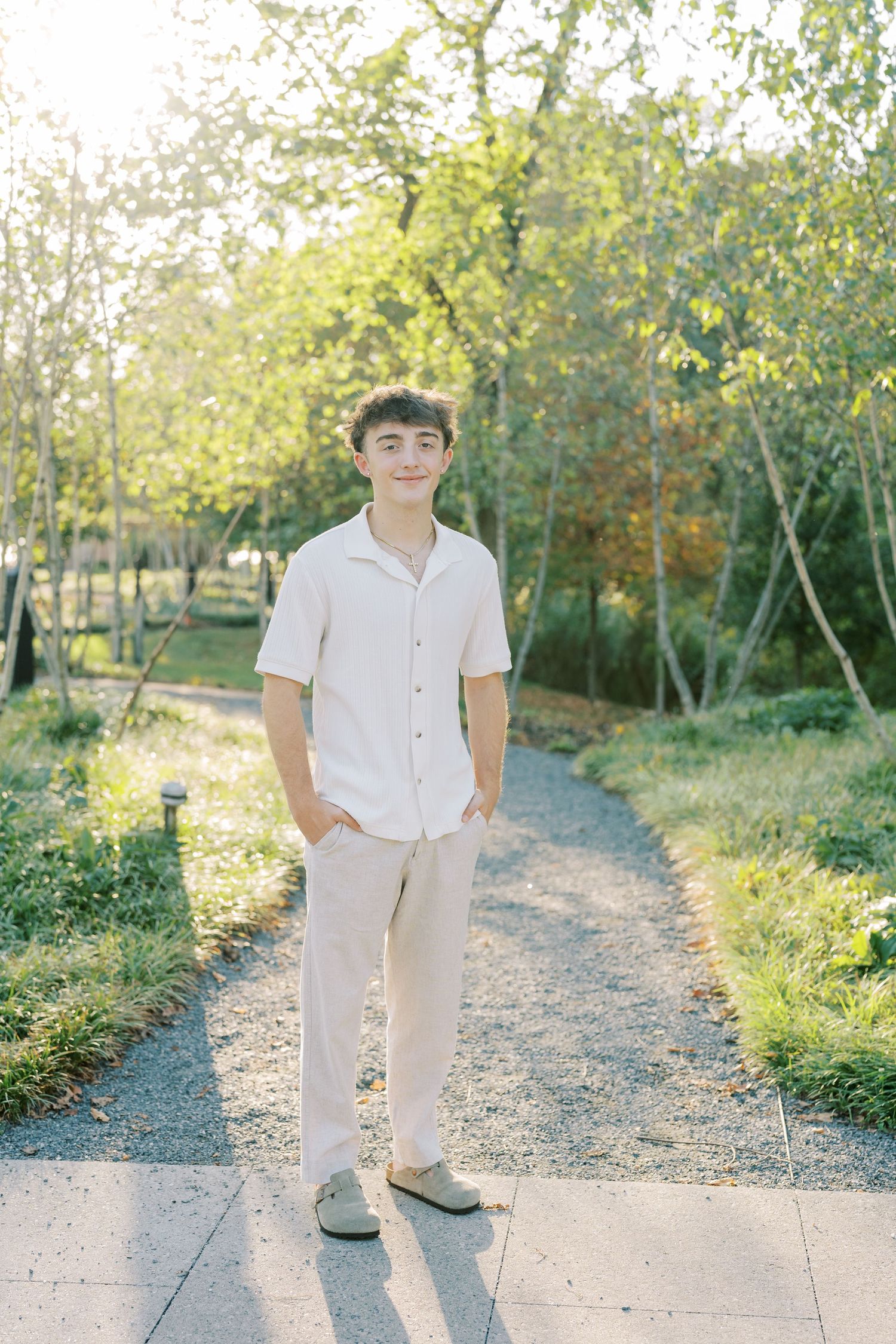Someone in all-white casual attire stands on a gravel path surrounded by lush green trees on a sunny day.