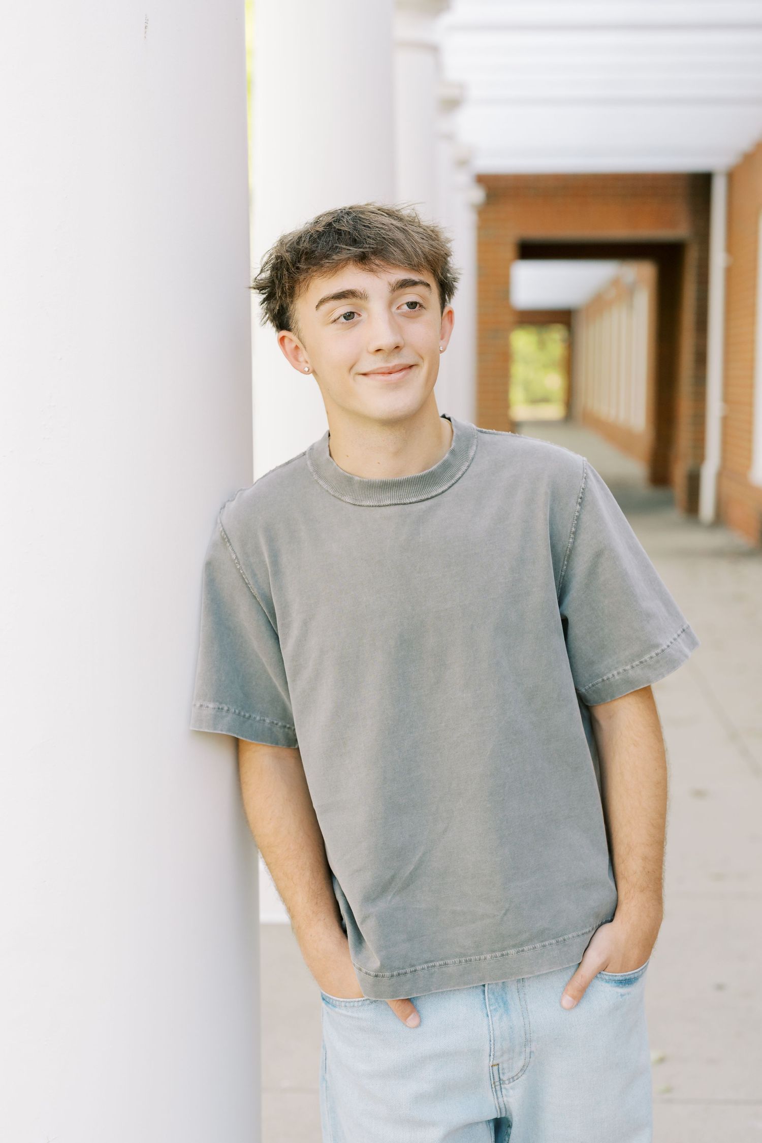 Person wearing a gray t-shirt and light blue jeans poses casually against white columns in a hallway.