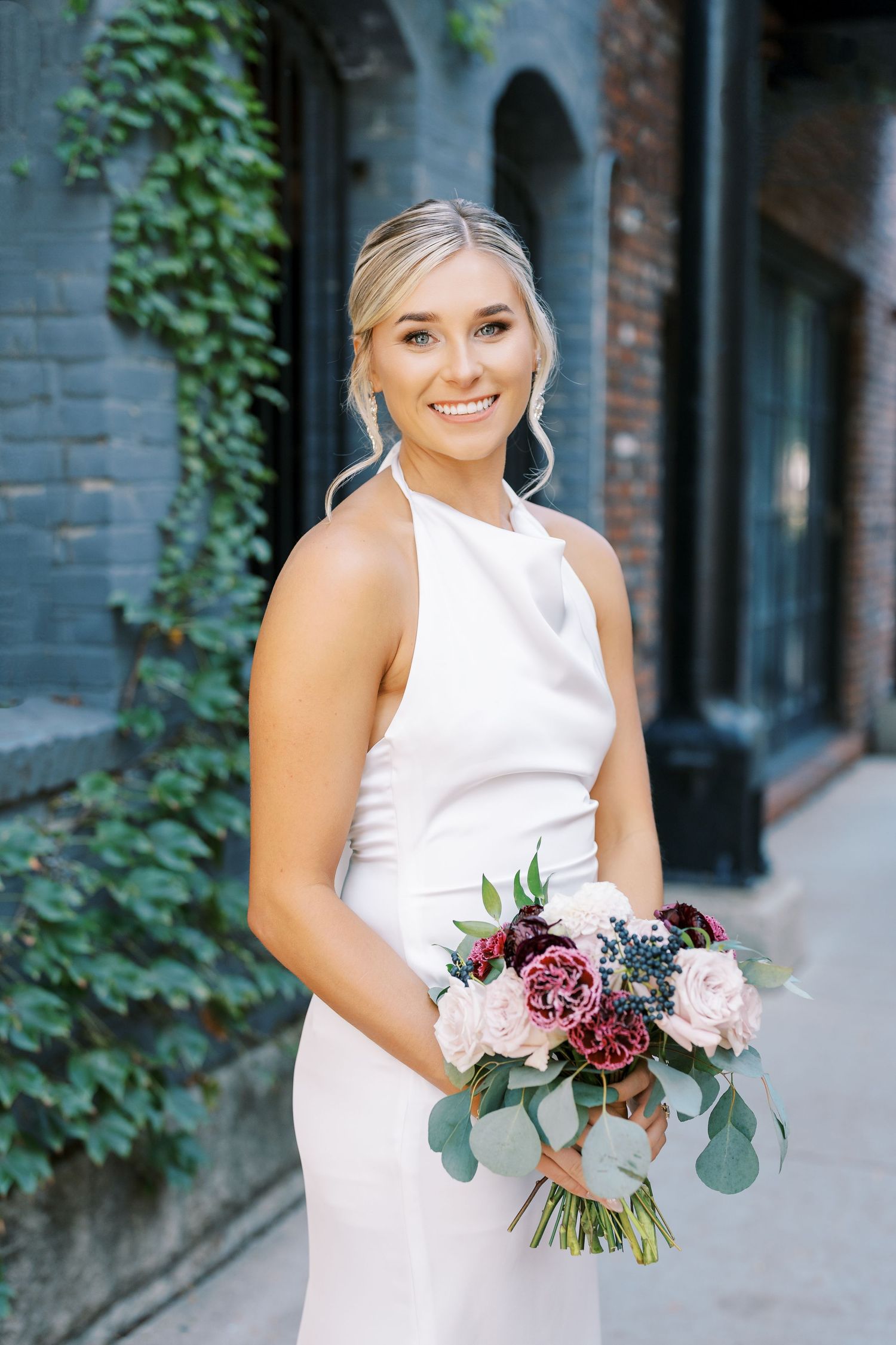 Elegant bride in white sleeveless dress holds romantic burgundy and blush bouquet with eucalyptus against brick wall.