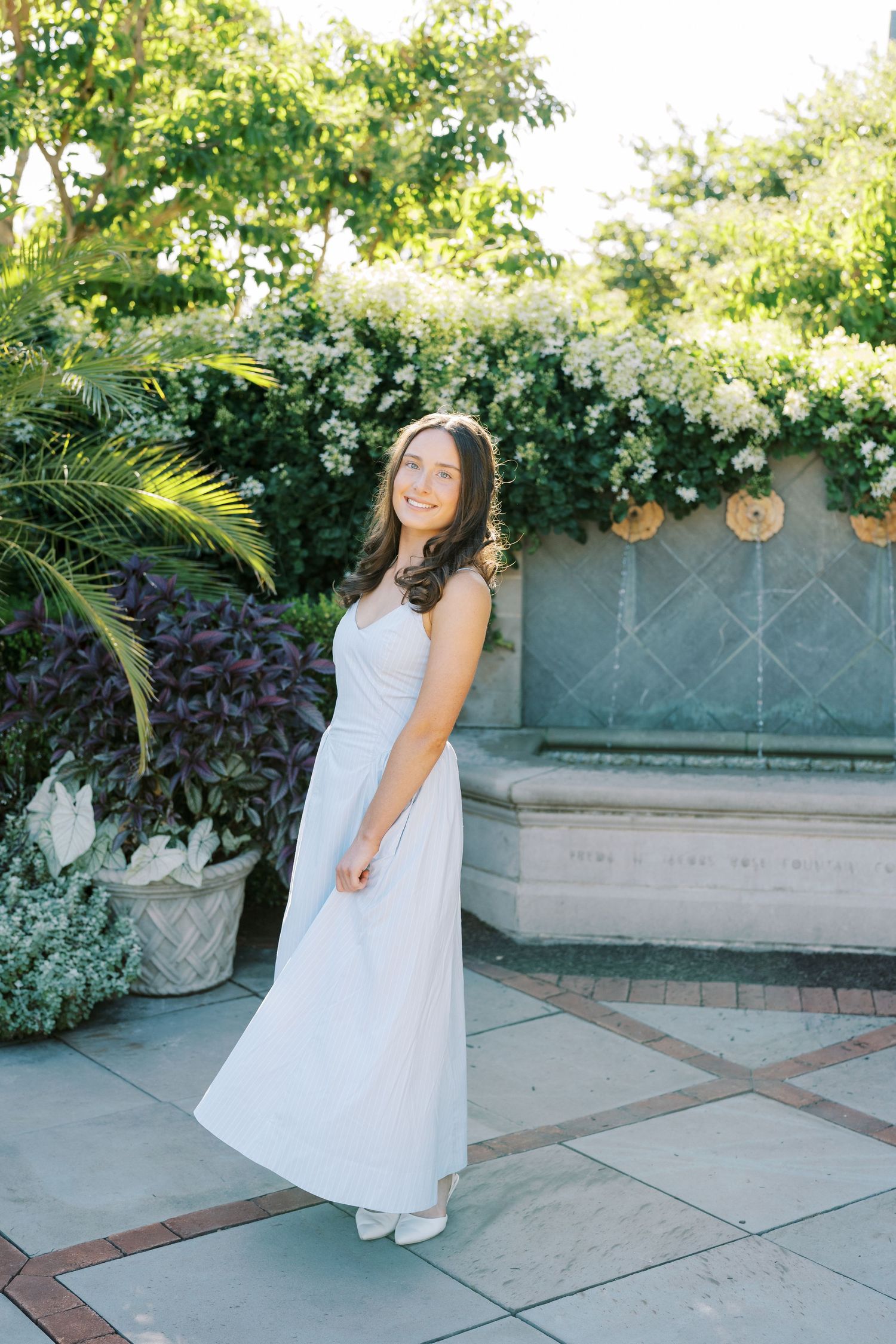 A person in a flowing white dress stands in a garden setting with lush greenery and decorative stone pavers.