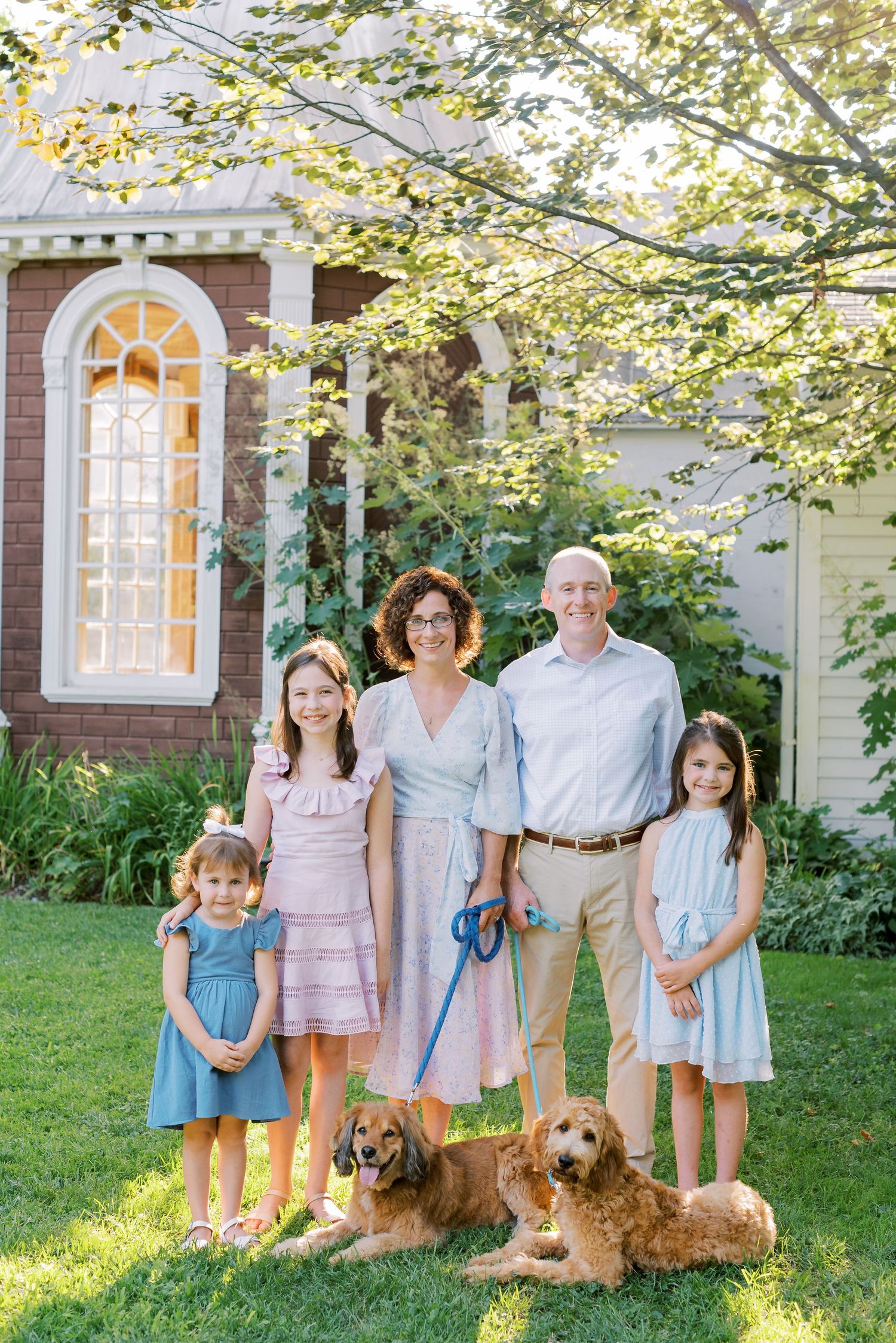 A family in pastel outfits poses with two golden doodle dogs on a sunny lawn with trees in the background.
