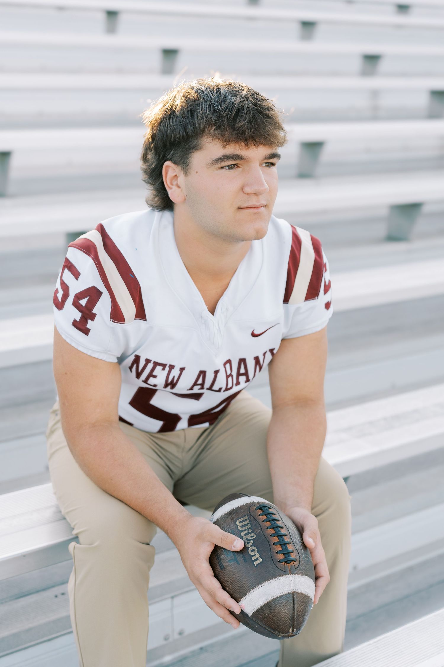 High school football player in white and maroon uniform poses on stadium bleachers with football.