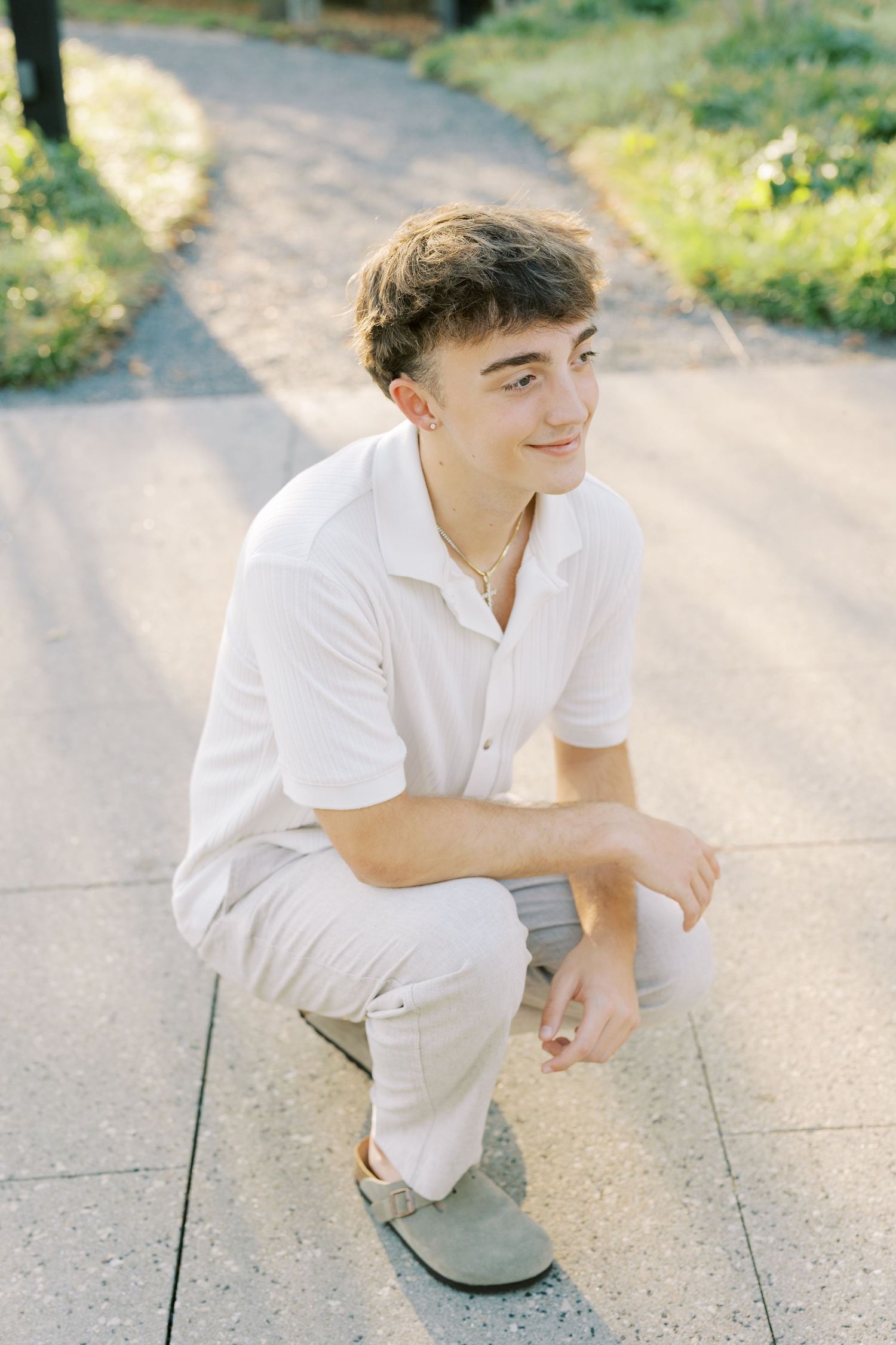 A person in white clothing crouches on a sunlit path while smiling and looking to the side.
