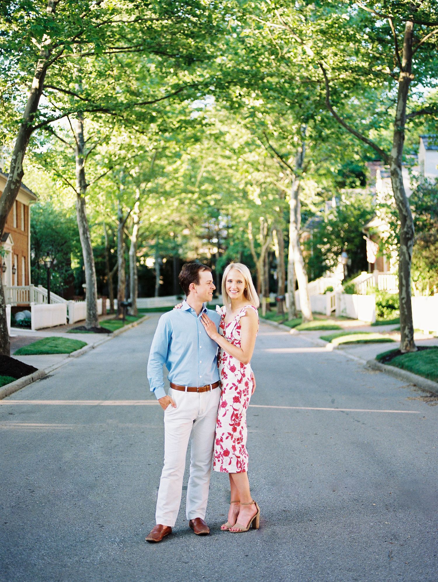 A couple walks together on a tree-lined street during a sunny engagement photo session.