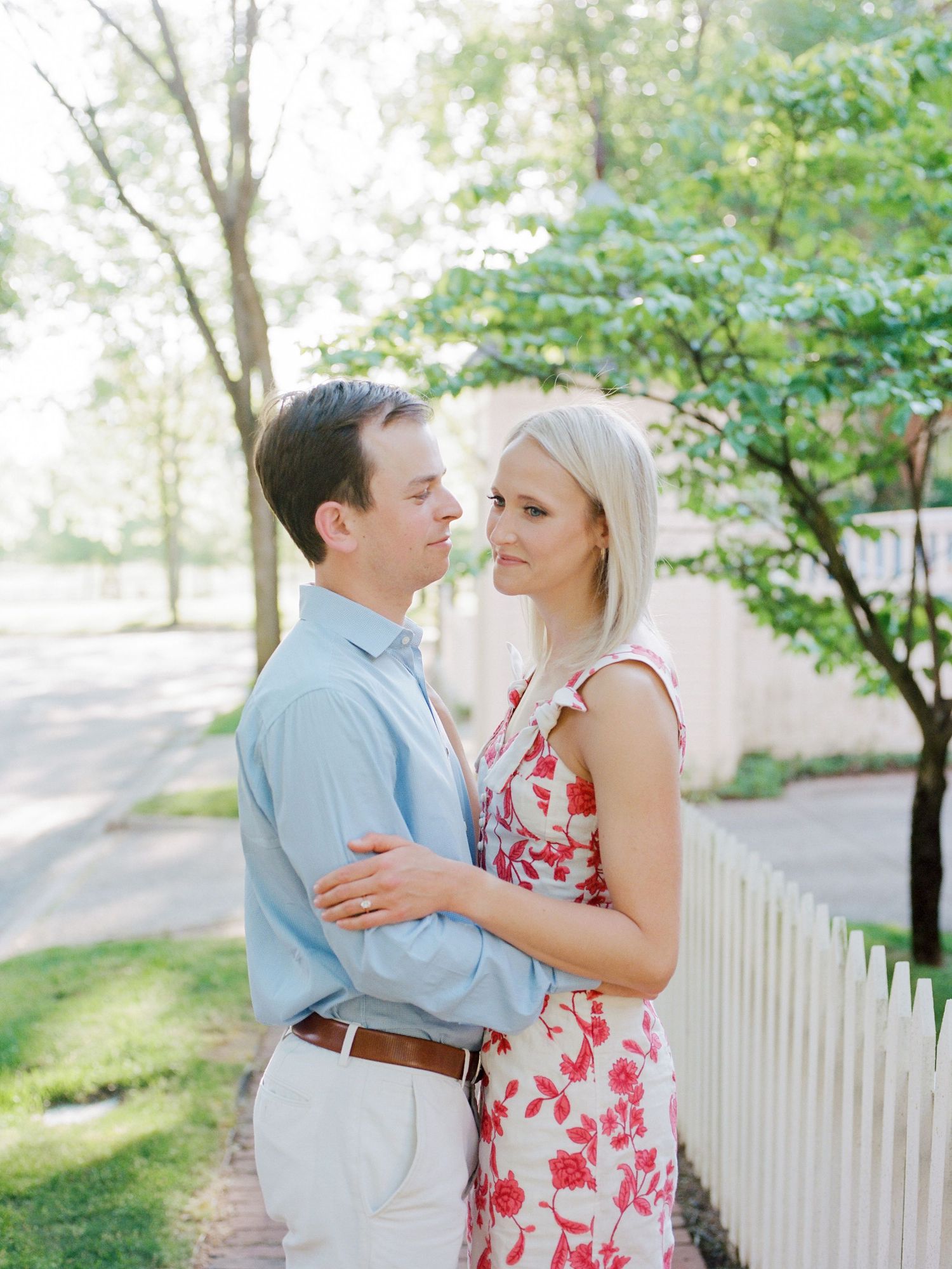 A couple shares a tender moment outdoors near a white picket fence on a bright summer day.