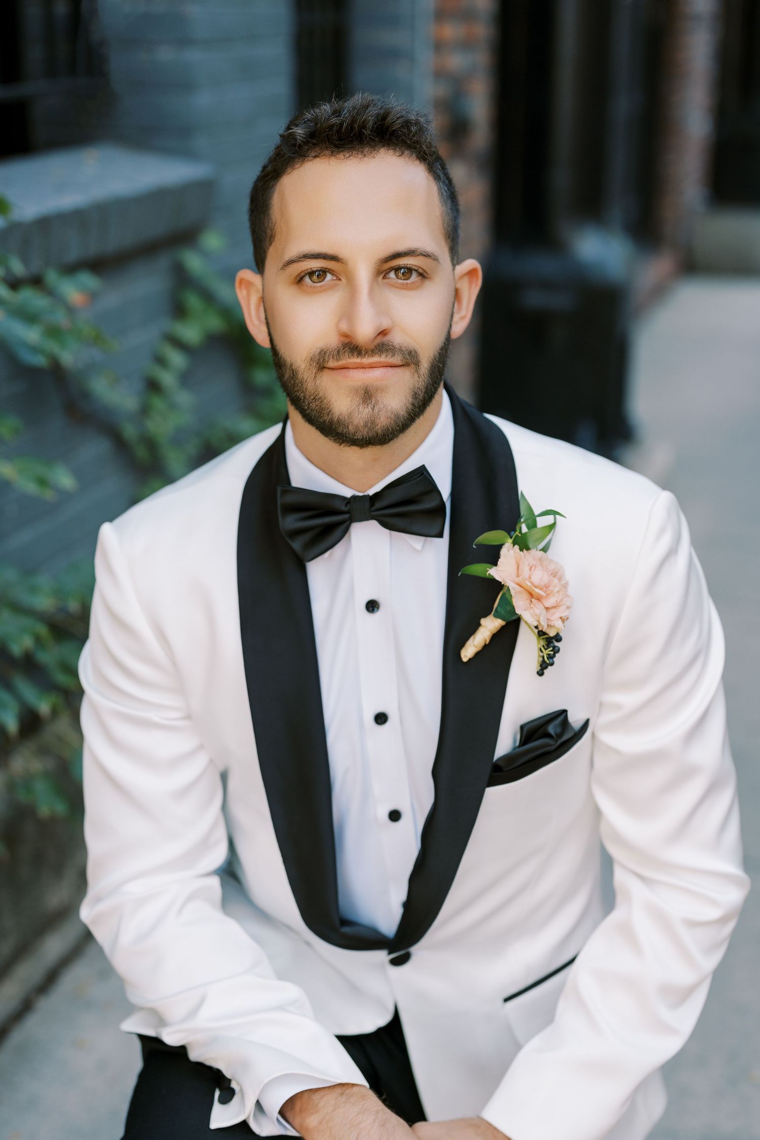 A groom in a white tuxedo jacket with black lapels and bow tie smiles at the camera in a formal portrait series.