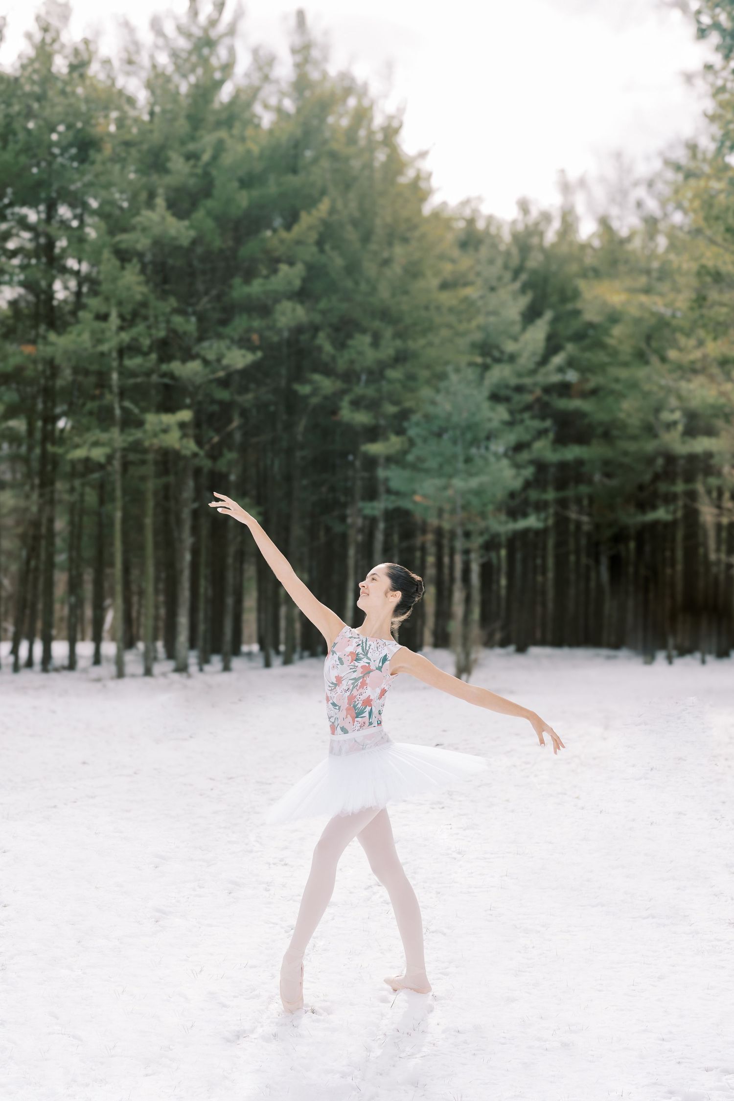 A dancer performs graceful movements in a snowy forest setting with evergreen trees creating a serene winter backdrop.