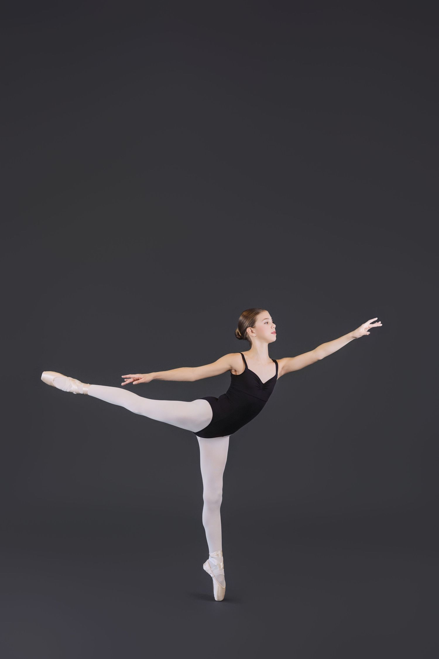 Ballet dancer in black leotard and white tights performs graceful arabesque pose against dark background.